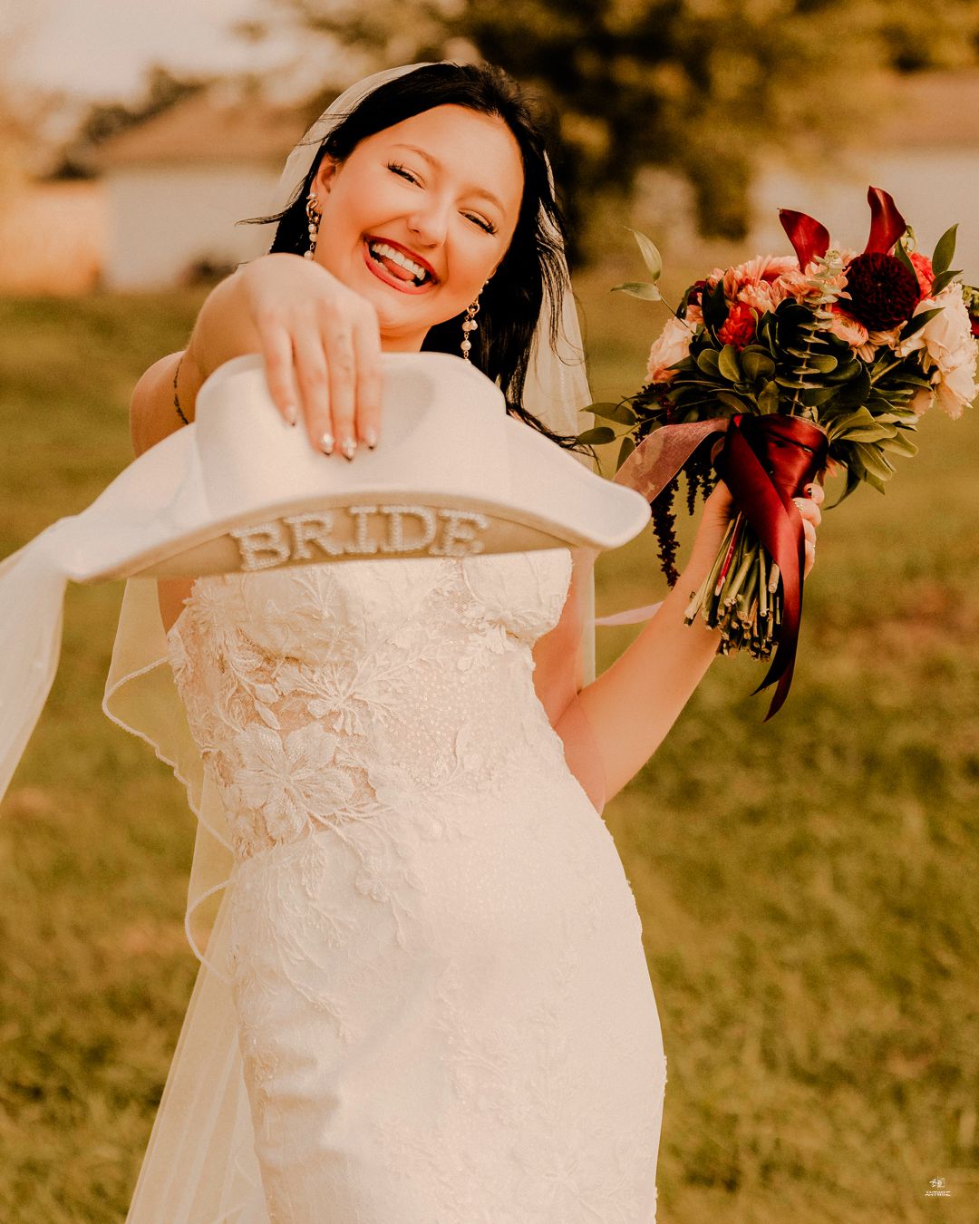 Smiling bride in strapless lace gown holding a white cowboy hat labeled BRIDE and floral bouquet.
