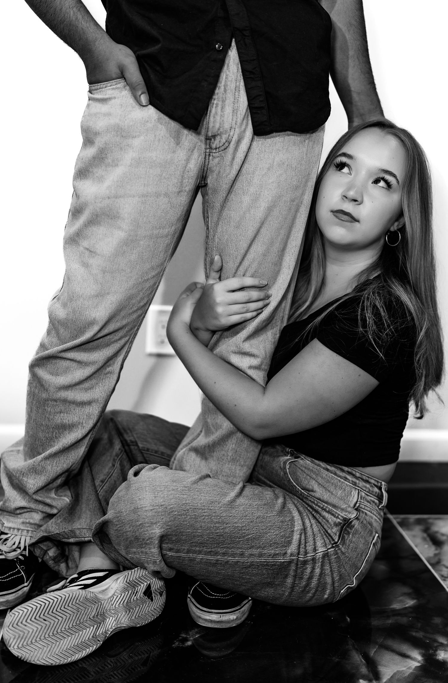Black and white studio portrait of a young woman sitting on the floor embracing a standing mans leg.