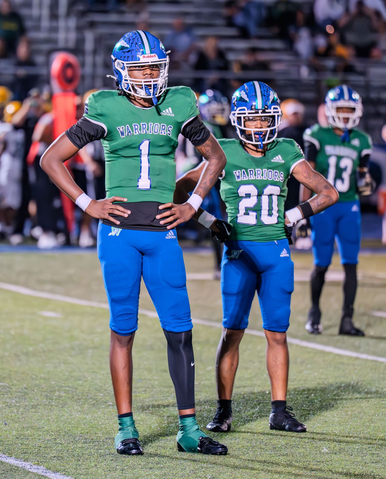 Warriors football players numbers 1 and 20 in green and blue uniforms stand together on the field.