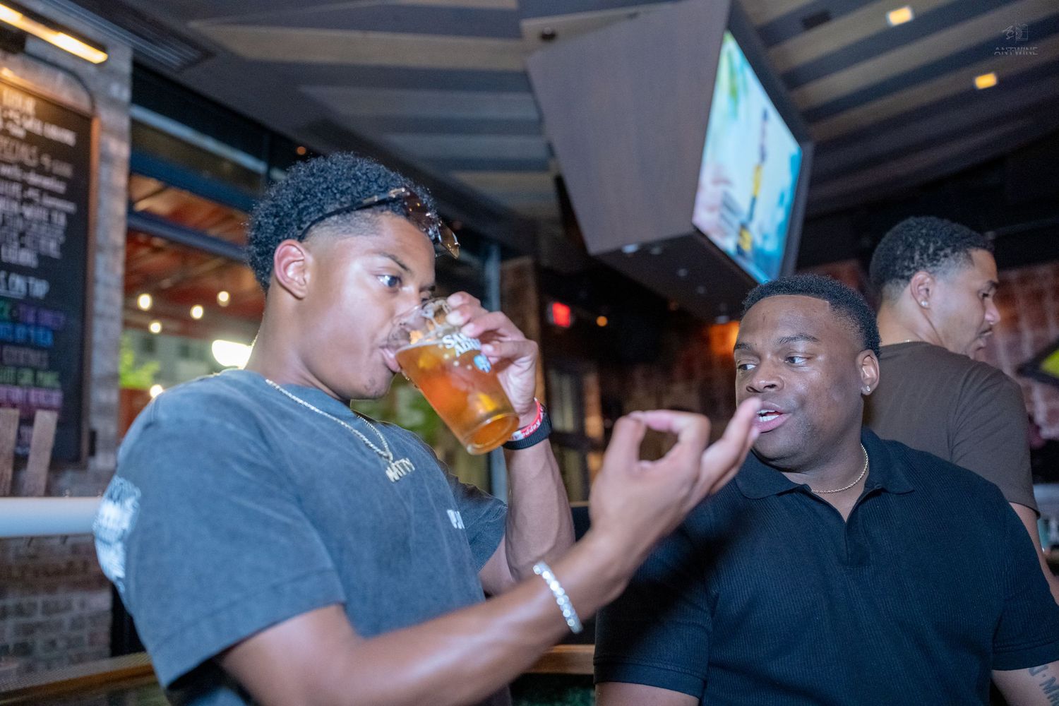 Two young men at a bar, one drinking a beer while the other snacks nearby.
