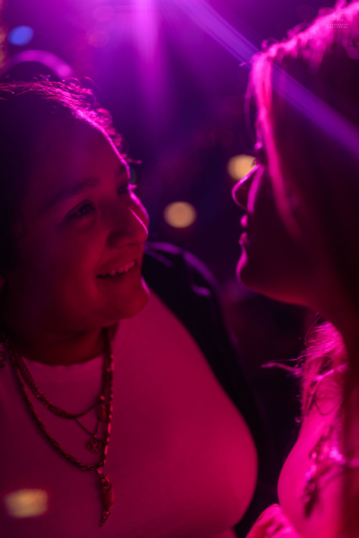 Two women facing each other smiling in vibrant pink and purple nightclub lighting.