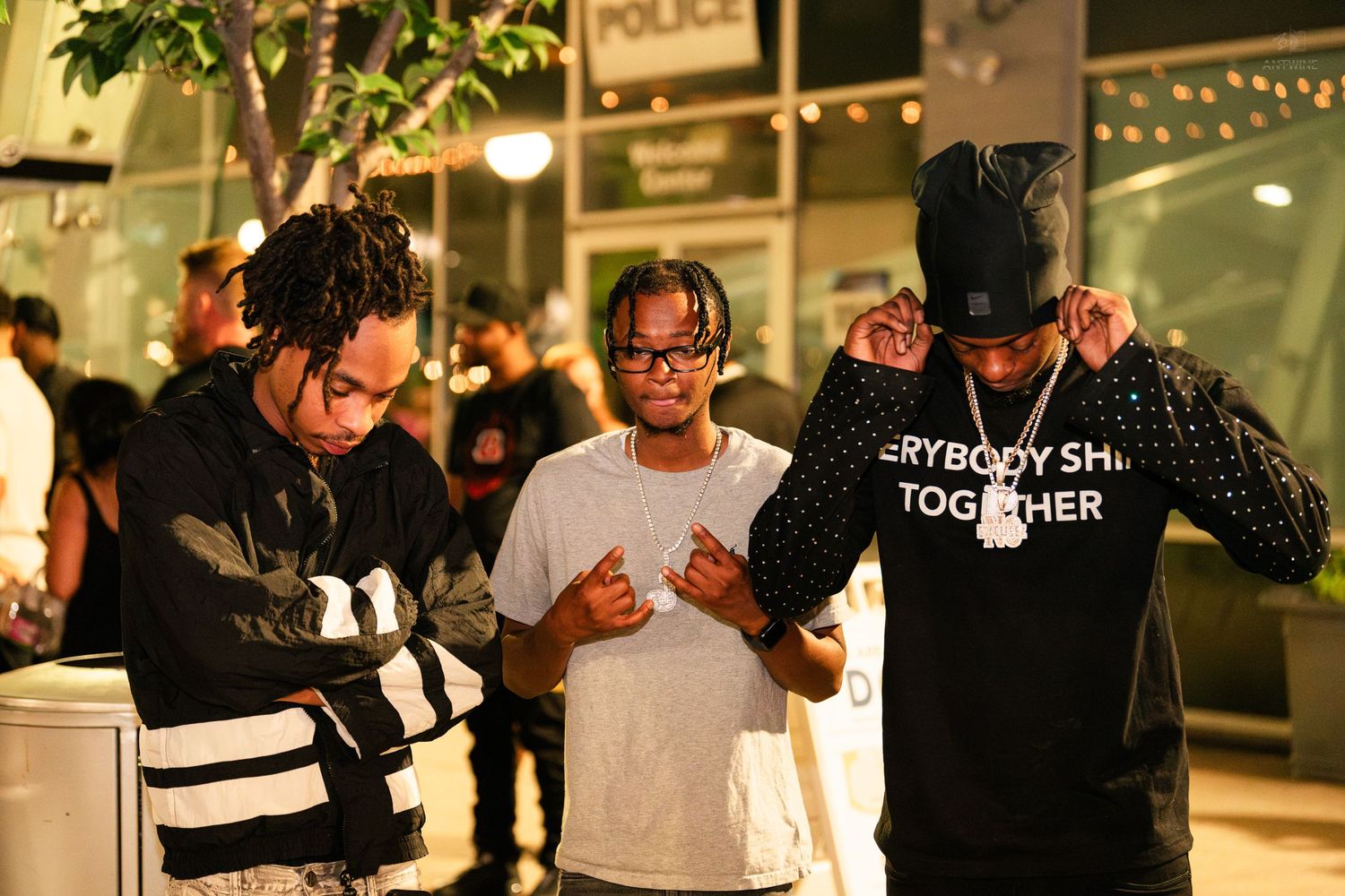 Three men in dark streetwear gather outside a building at night under bright outdoor lighting.