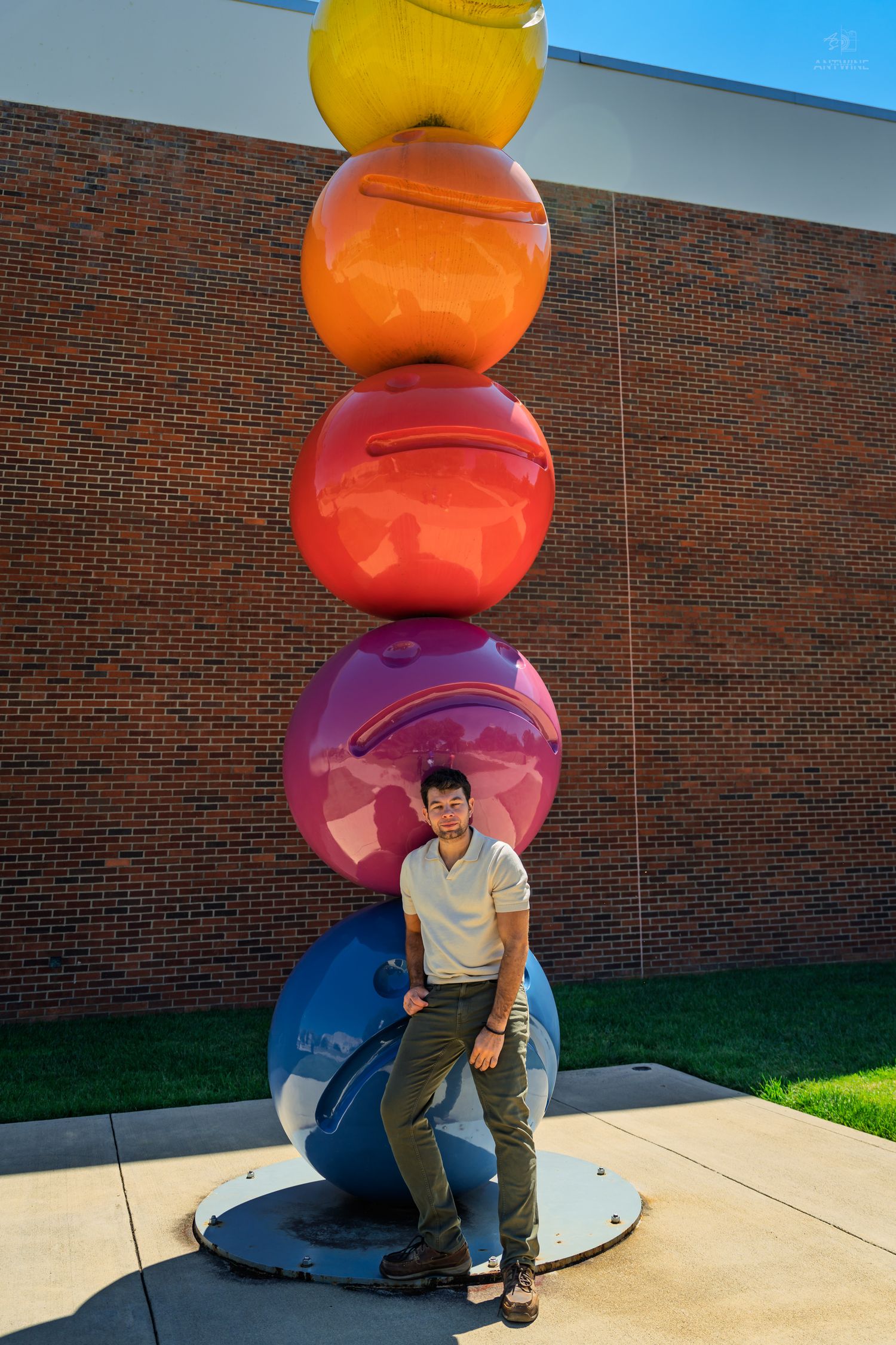Young man leaning against large multicolored stacked spheres public art sculpture outdoors.
