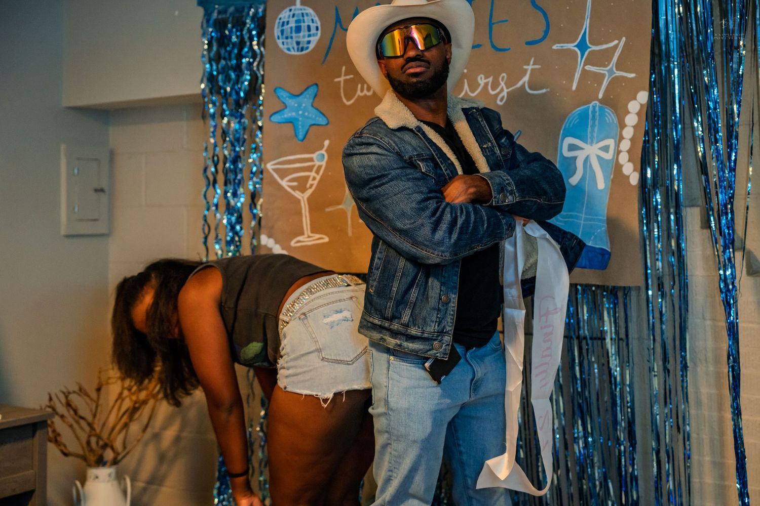 Man in cowboy hat and denim jacket poses at blue-themed birthday party backdrop.