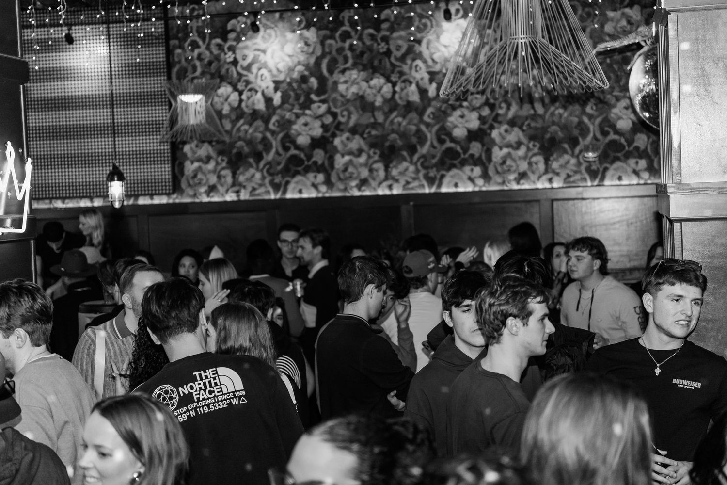 Black and white wide shot of a packed nightclub crowd under decorative lights and floral wallpaper.