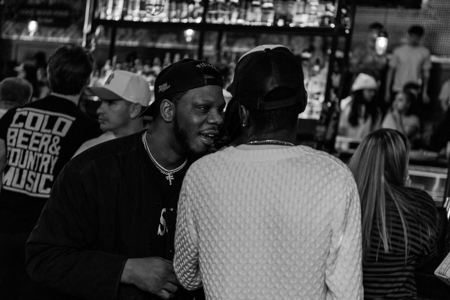 Two men having a conversation near a bar in a nightclub setting.