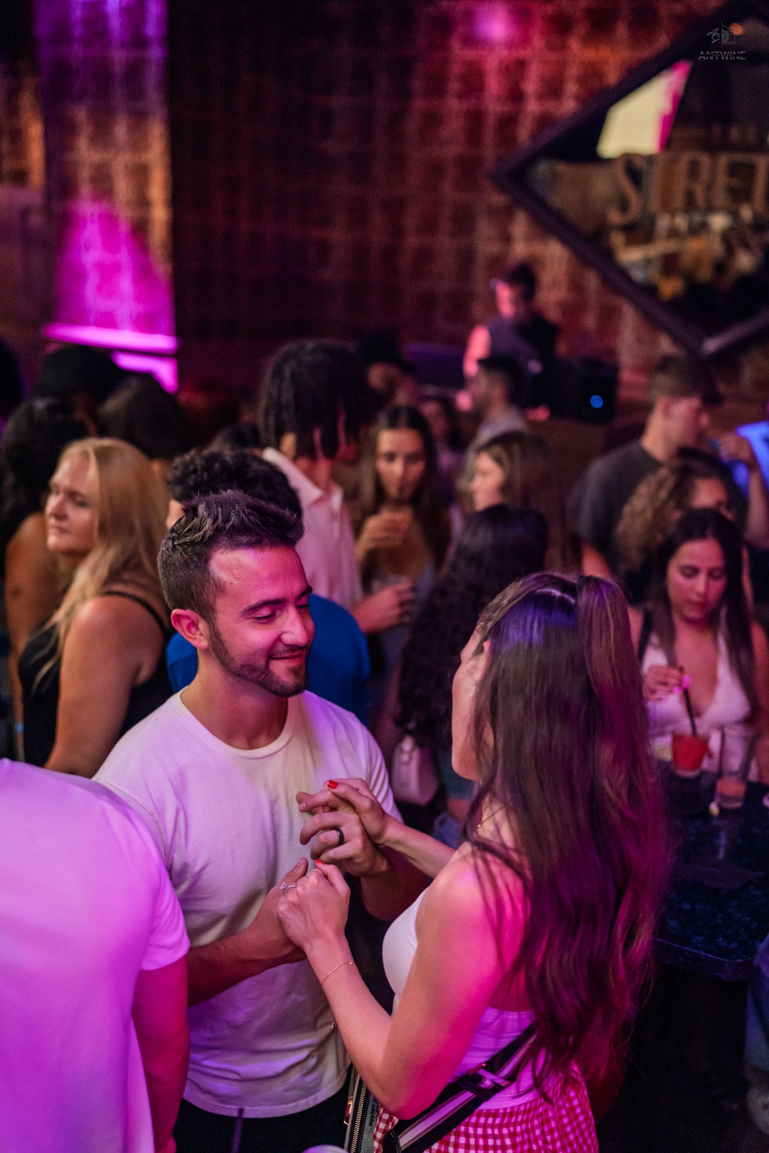 Couple holding hands and smiling at each other on a vibrant pink-lit nightclub dance floor.