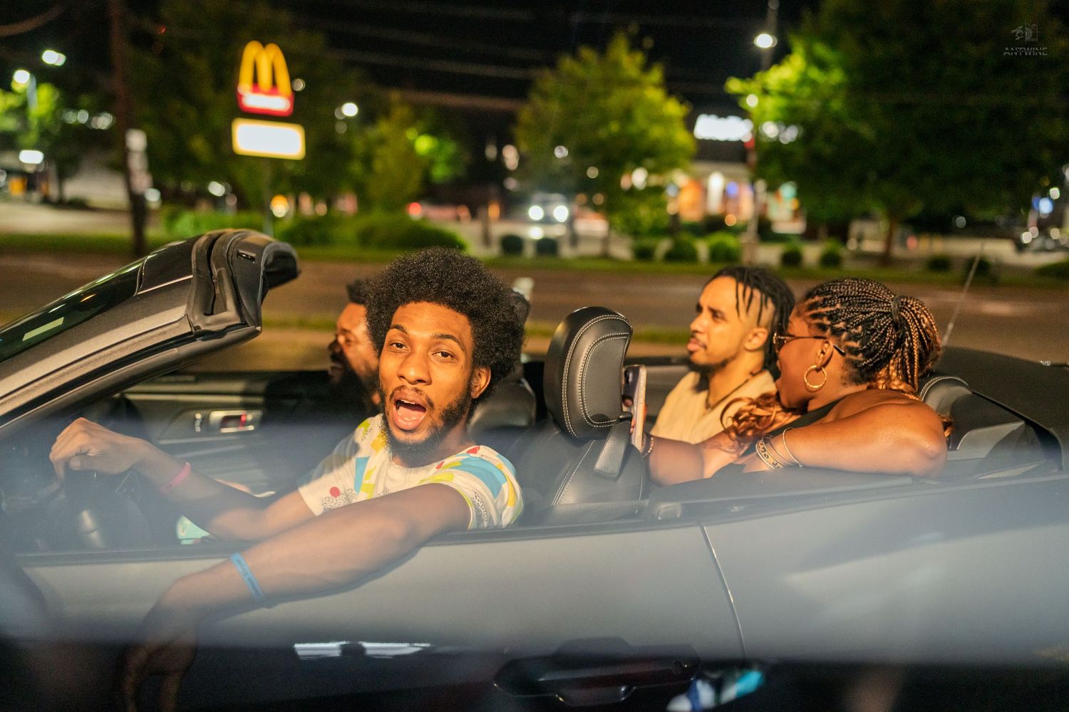 Four friends laughing and having fun in a convertible car at night near a McDonald's.