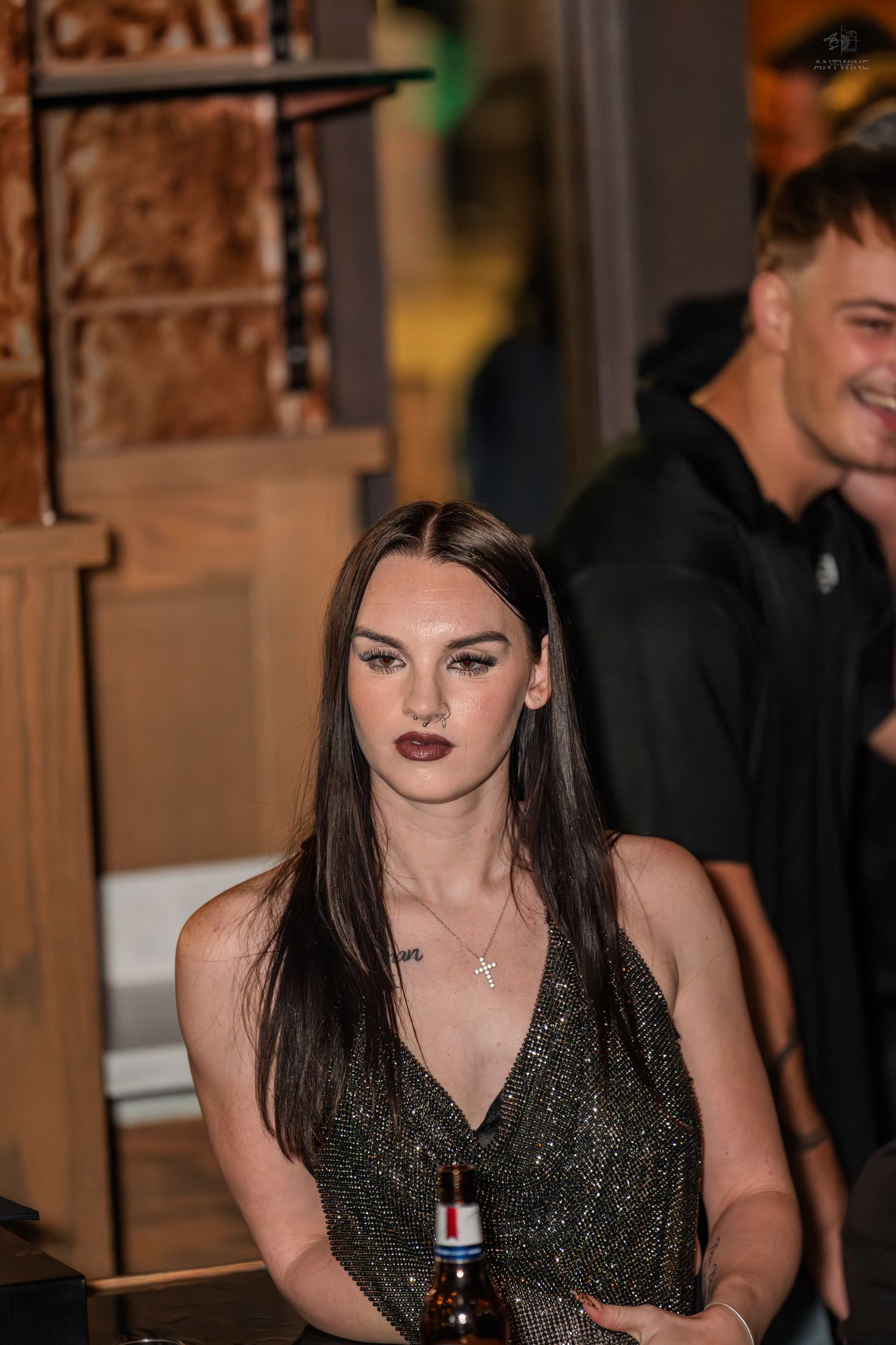 Young woman with dark hair in sparkly dress holding beer at bar.