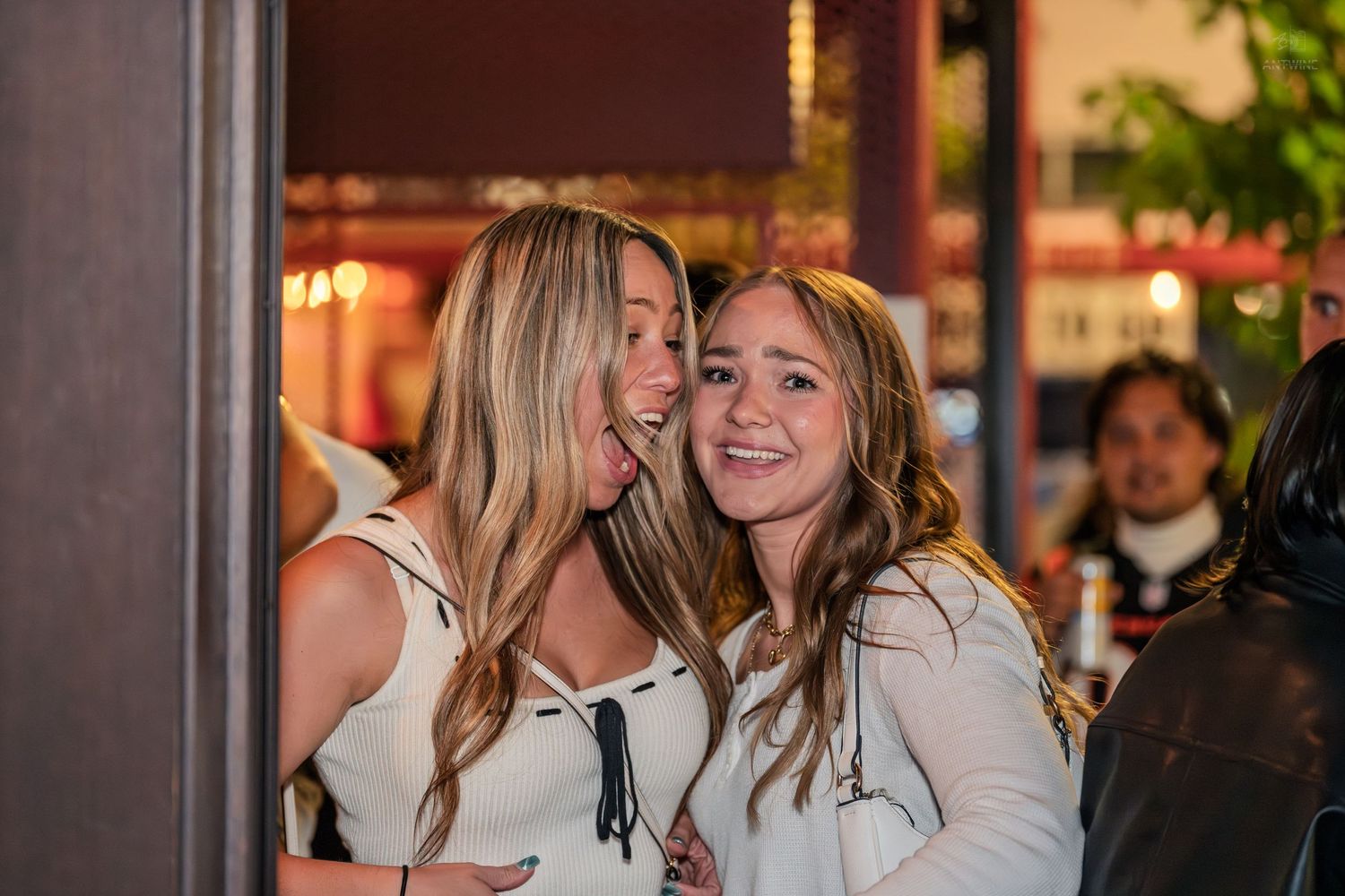 Two young women in white tops share a joyful embrace at an evening social gathering.