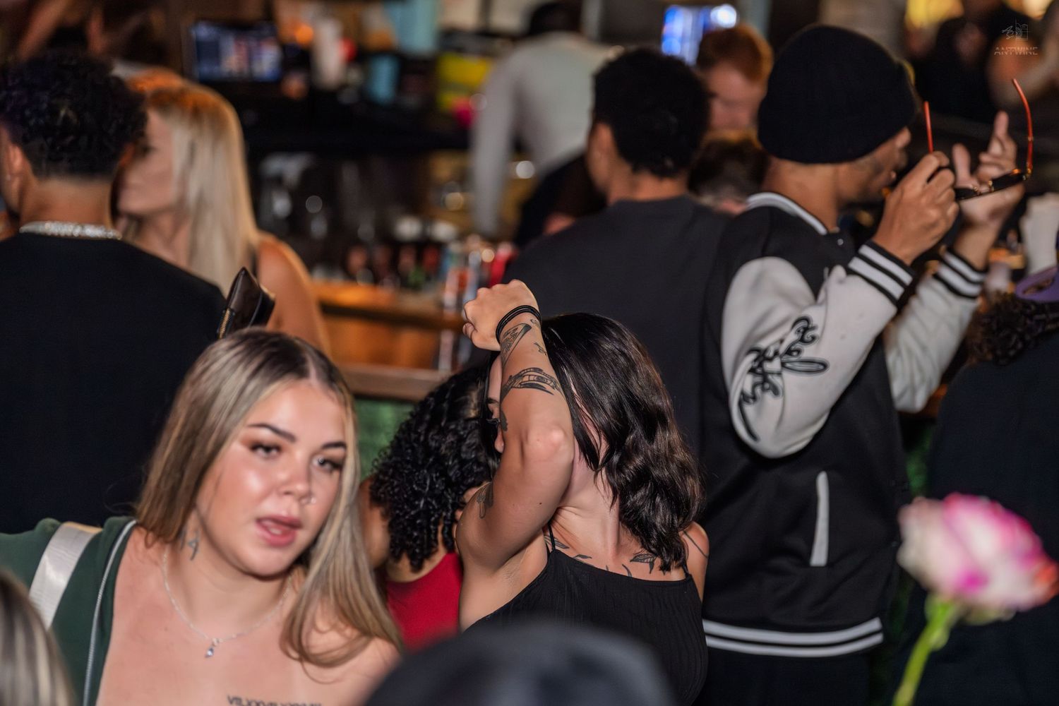 Crowded nightclub bar scene with diverse group of young adults socializing under warm ambient lighting.