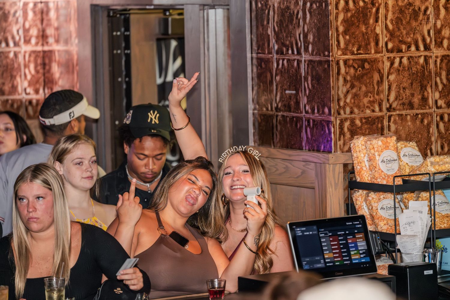 Cheerful group of friends partying at a bar, woman in tiara smiling with phone.