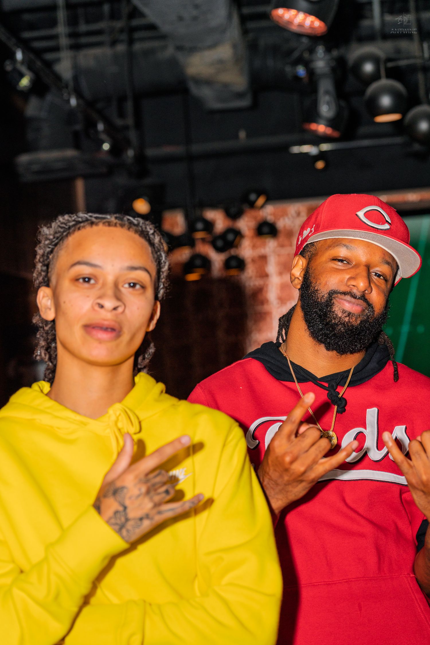 Man in red Cincinnati Reds jersey and woman in yellow hoodie posing together at nightclub event.