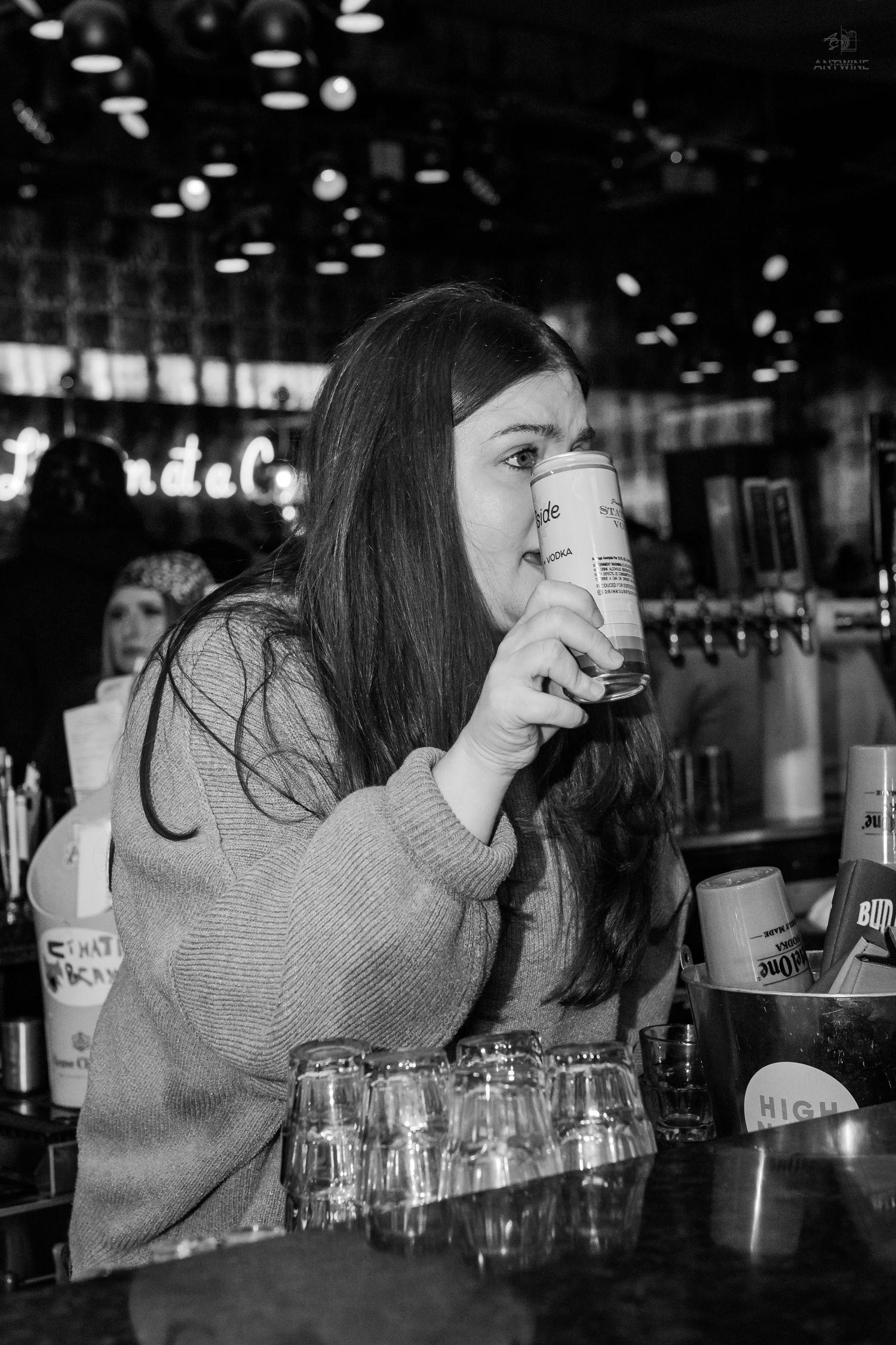 Black and white photo of a young woman drinking from a can at a bar.