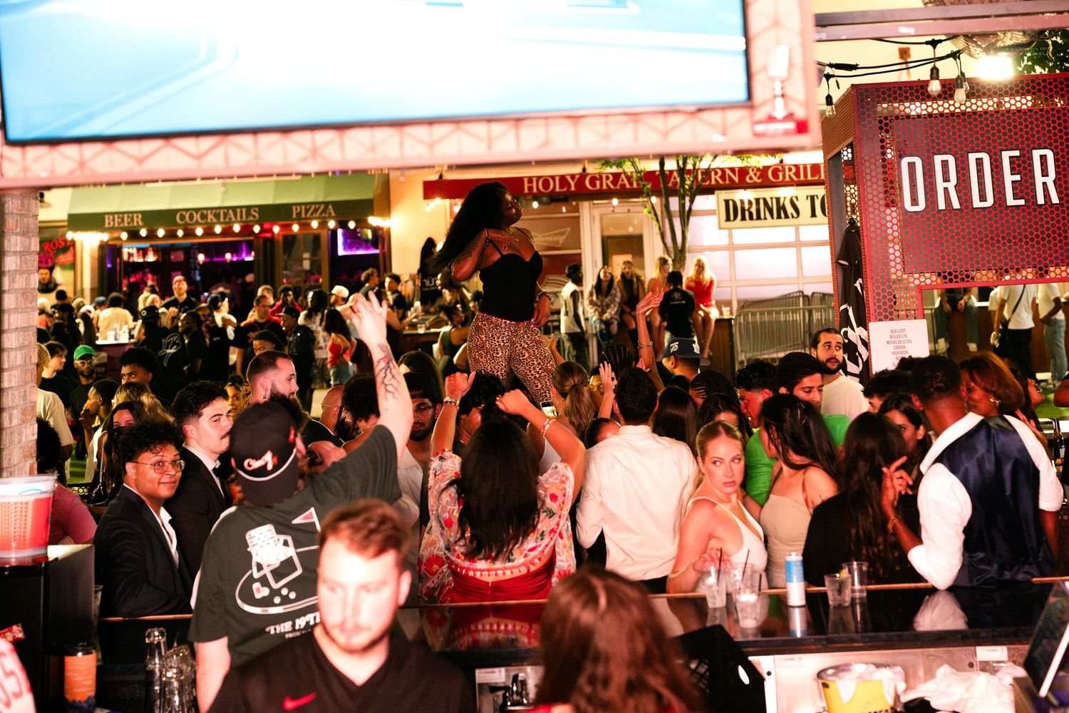 Energetic crowd dancing and celebrating at a packed nightclub bar with green lighting.