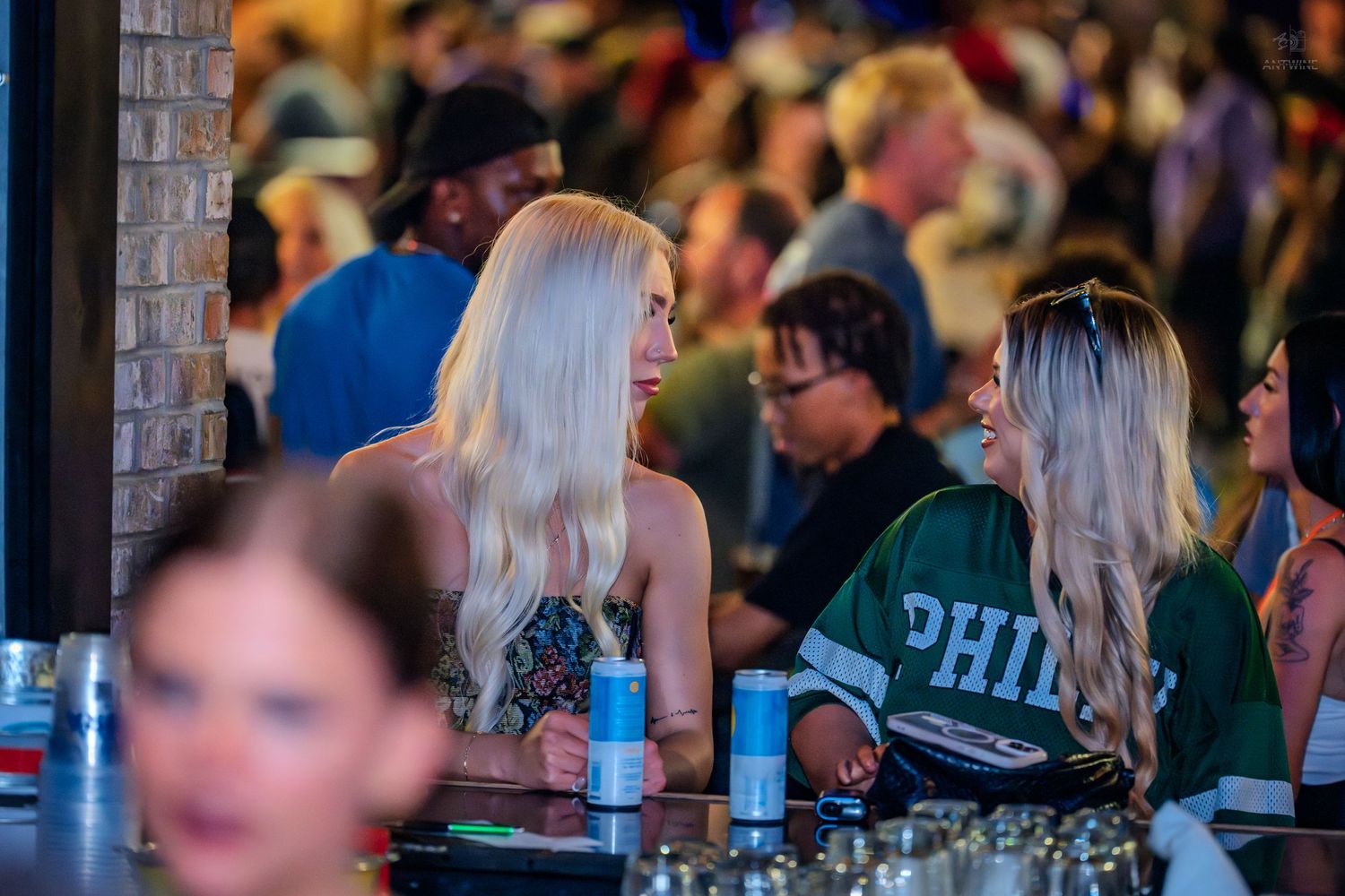 Two blonde women chatting at a bar with a lively crowd behind them.