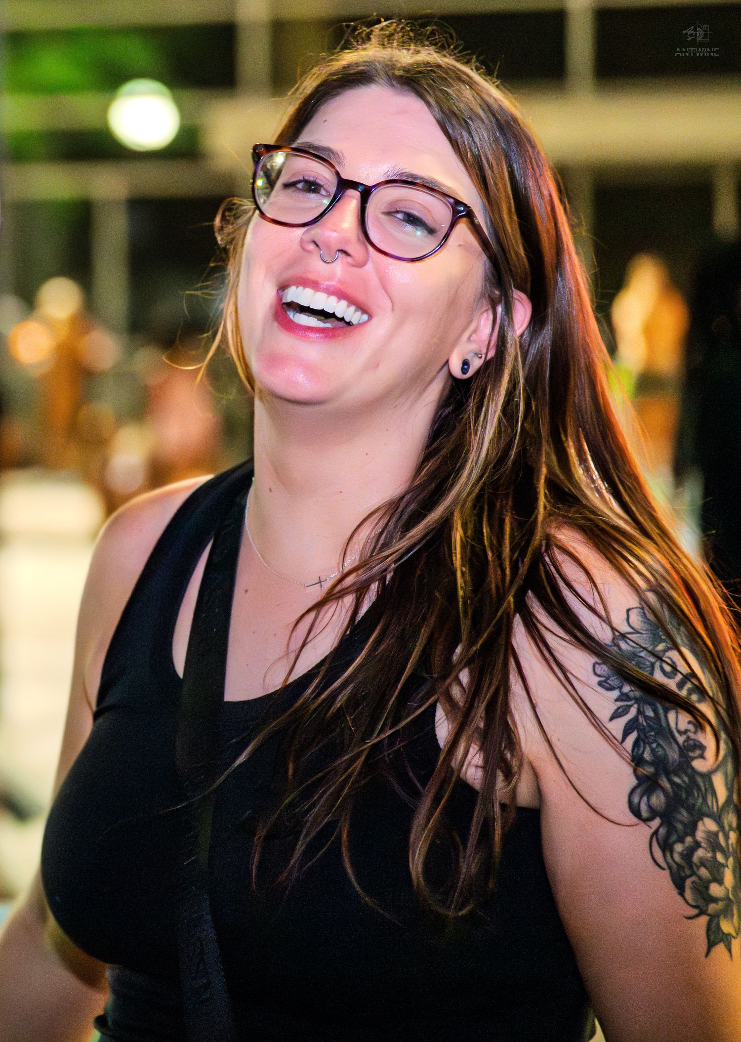 A laughing woman with long brown hair, glasses, and a floral shoulder tattoo in a black tank top.