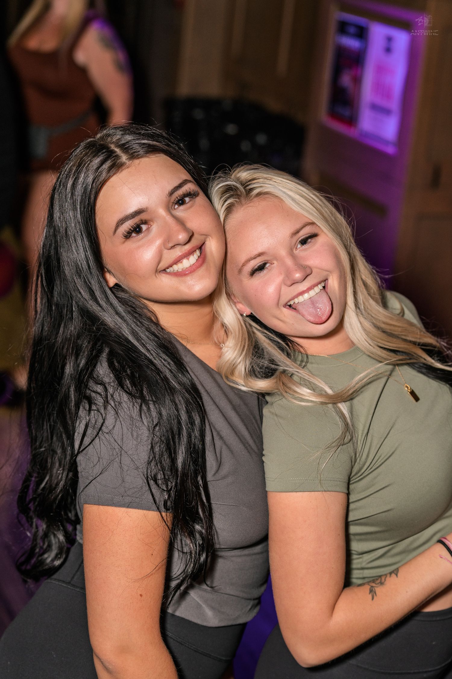 Two cheerful young women pose together at a nightclub, one smiling and one sticking out her tongue.