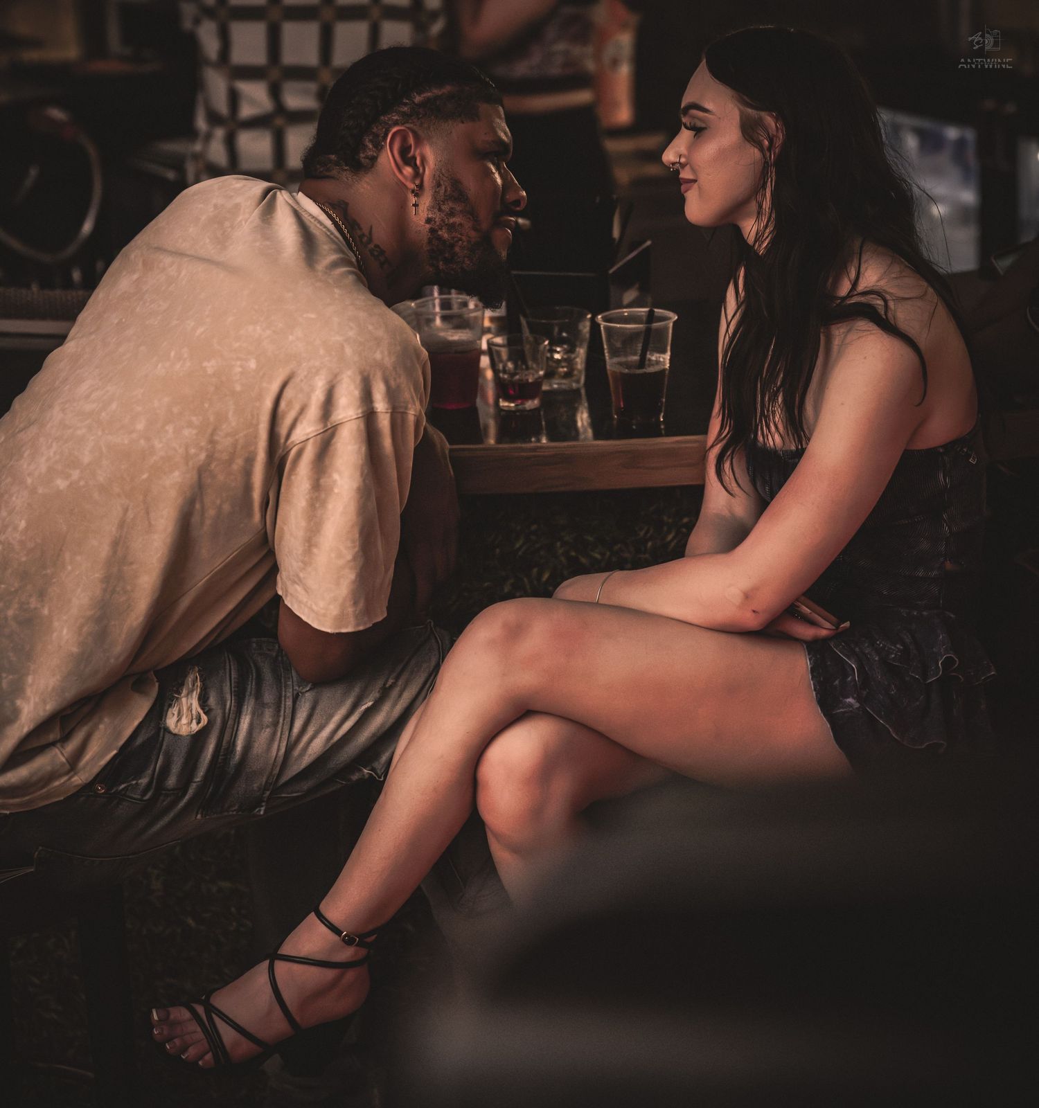 A man and woman share an intimate conversation at a dimly lit bar, gazing into each other's eyes.