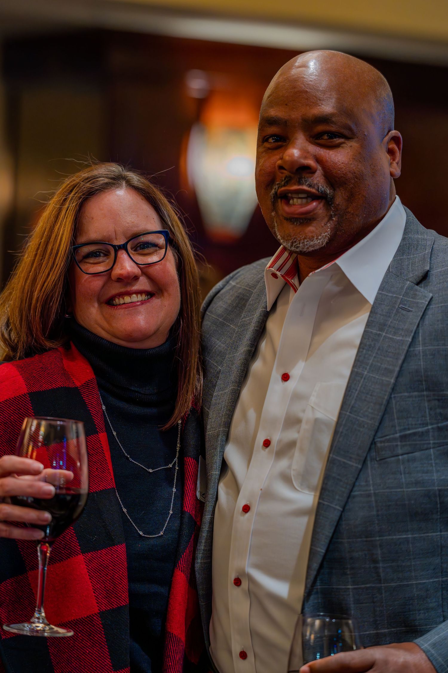 Two smiling adults holding wine glasses pose together at an indoor social event.