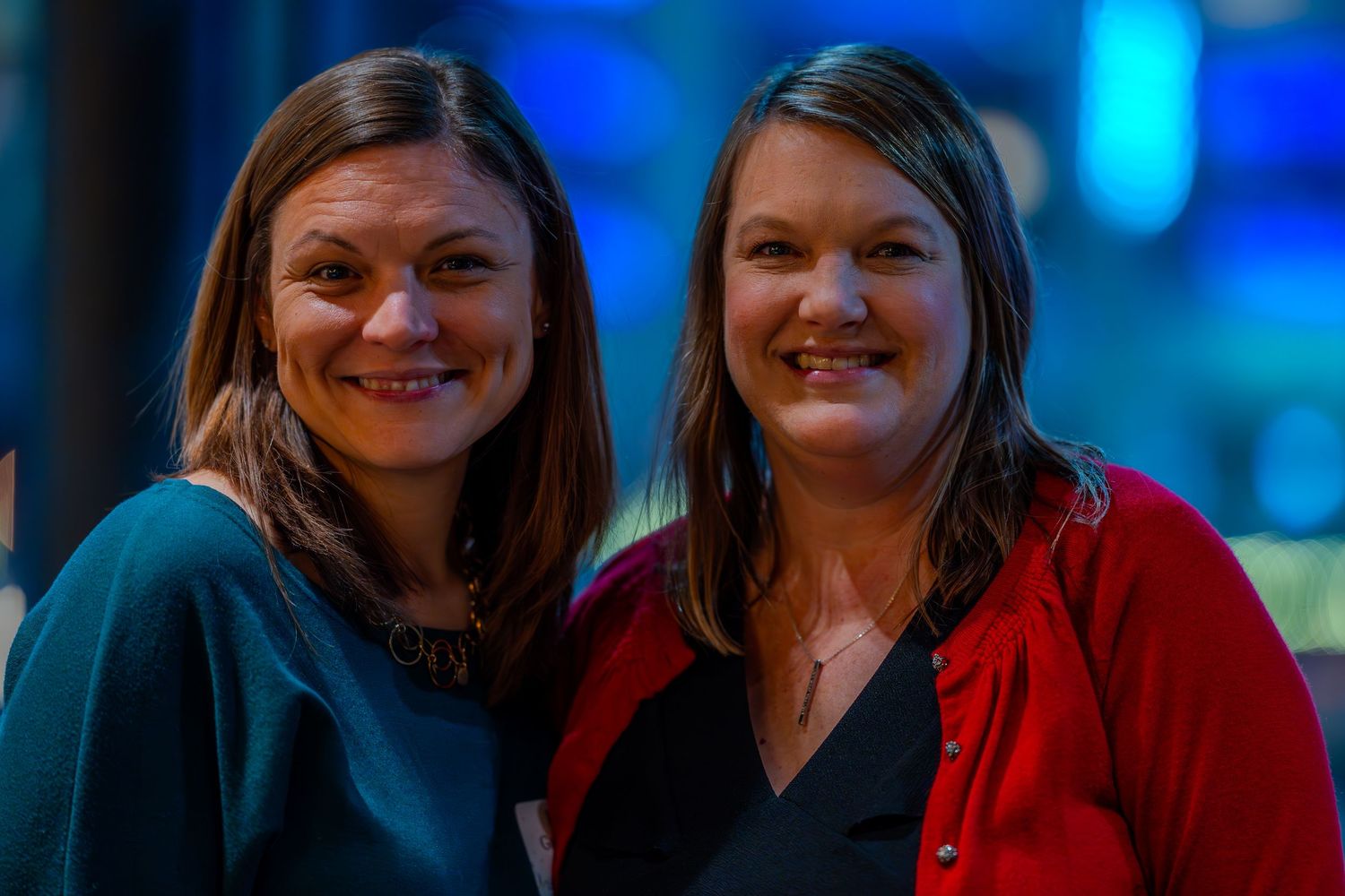 Two smiling women posing together indoors, one in teal and one in red cardigan, with colorful bokeh lights in background.