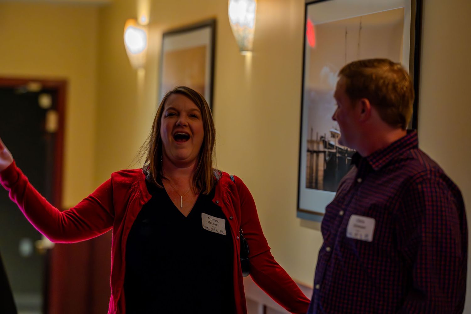 Woman in red cardigan laughing expressively with man in plaid shirt at indoor networking event.