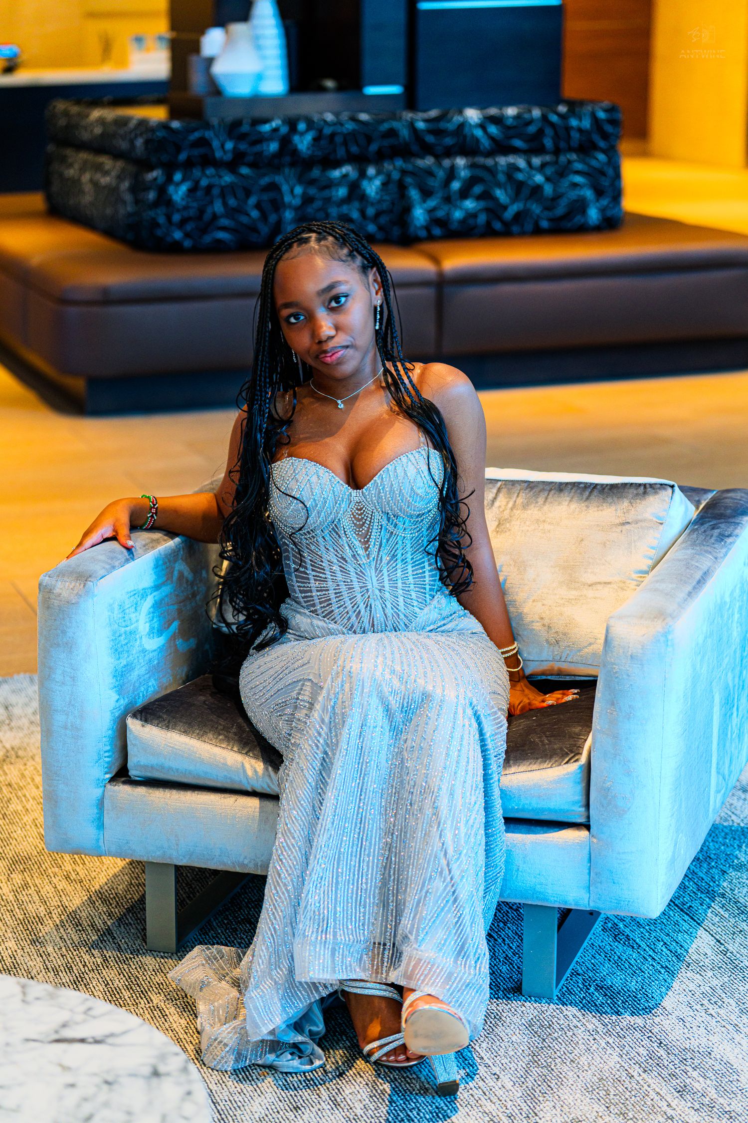 Young woman in silver sequin gown with braids poses elegantly in a velvet chair in a modern hotel lobby.