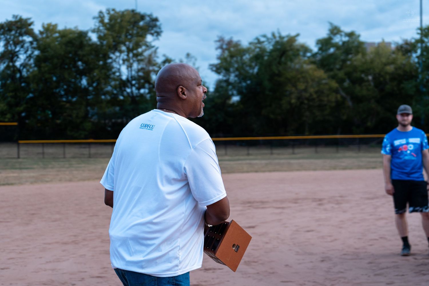 Man in white COPFCU shirt holds a clipboard on a softball field during a charity event.