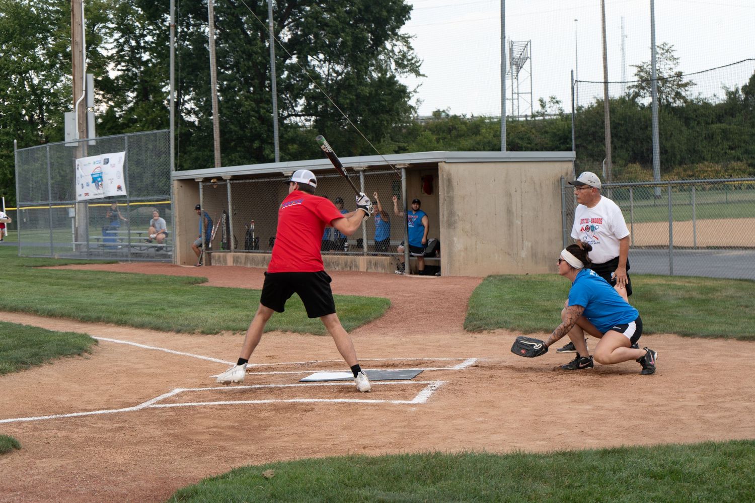 A batter in red swings at a softball pitch during an outdoor game, with catcher and umpire behind home plate.