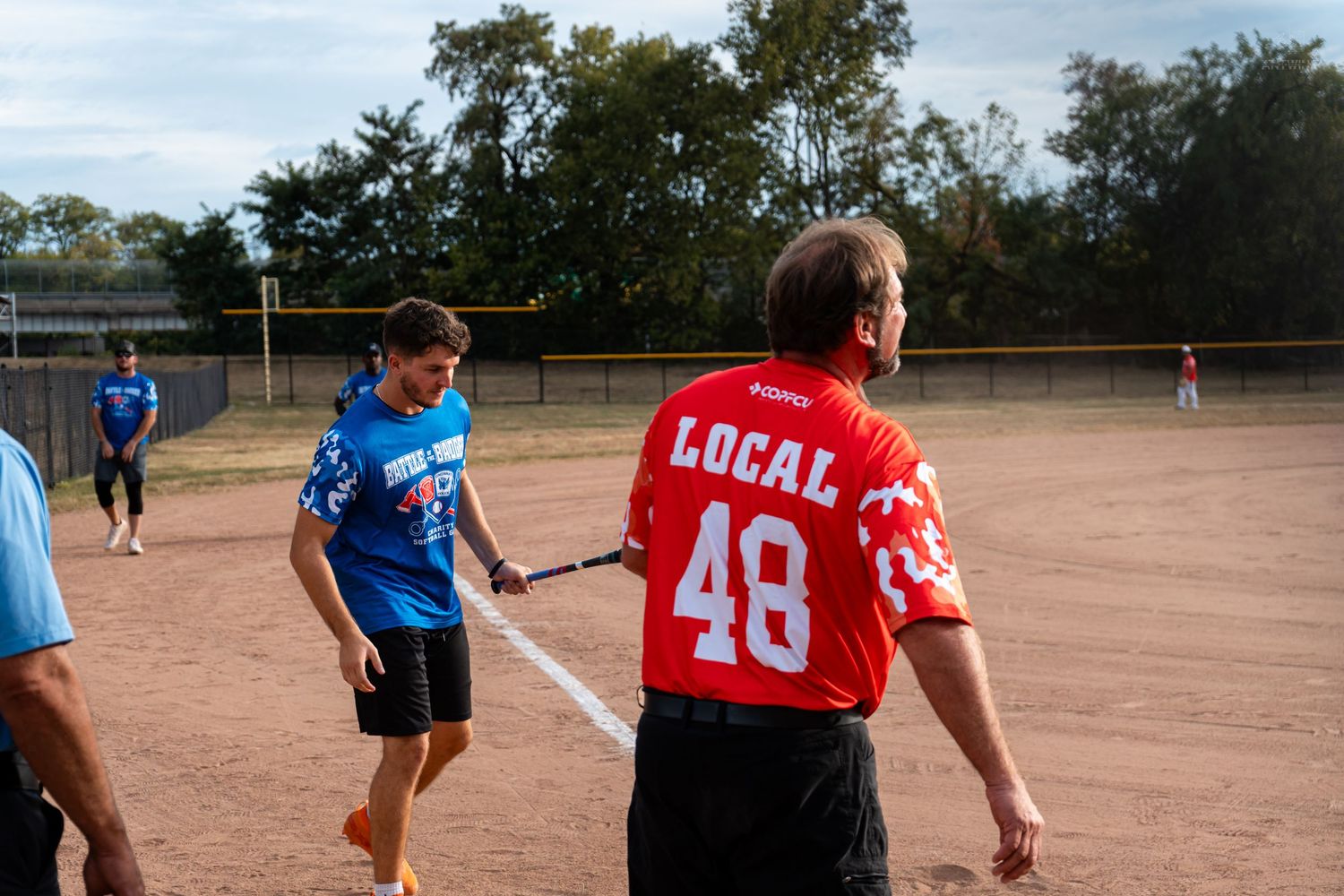 Two softball players in red Local 48 and blue jerseys shake hands on the dirt field after a game.