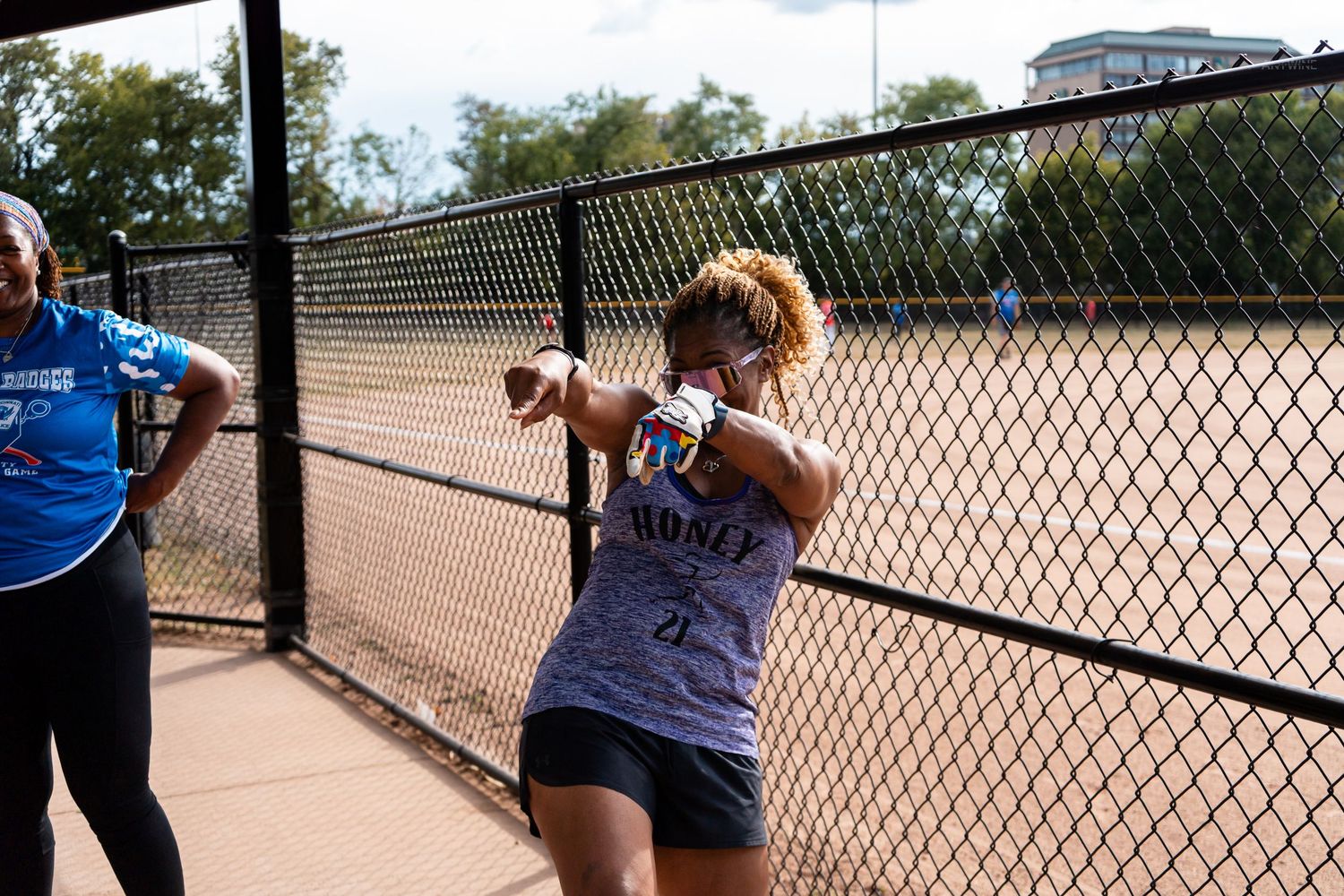 Woman in gray tank top stretches by chain-link fence at outdoor sports court on sunny day.