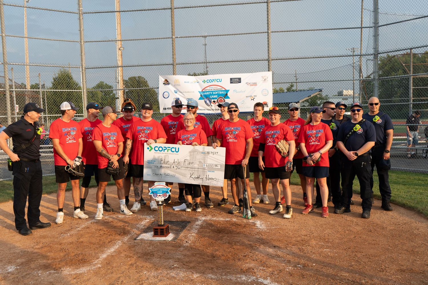 Softball team in red jerseys celebrates championship win holding oversized check on baseball diamond.