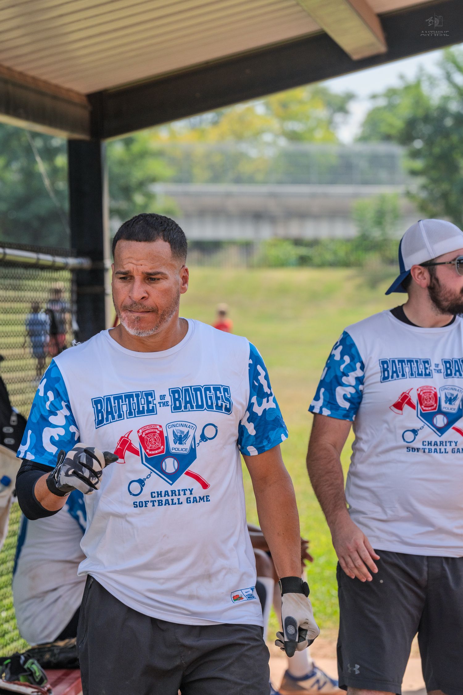 Two men in Battle of the Badges charity softball game jerseys stand in a dugout during an outdoor event.
