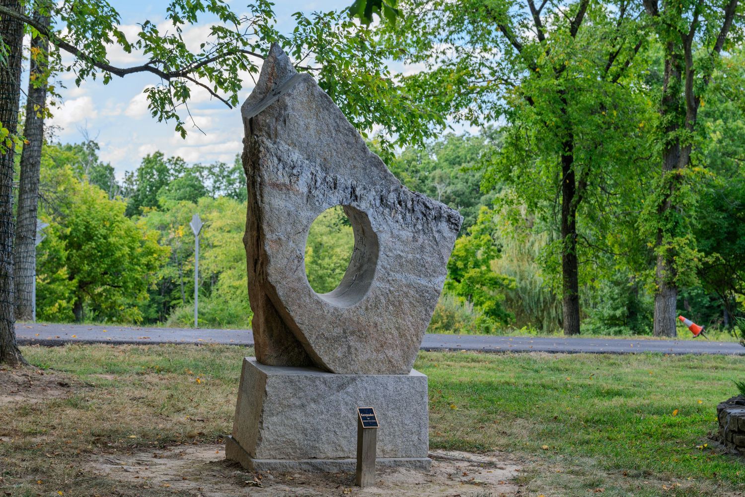 Large abstract stone sculpture with circular hole displayed in a lush green park setting on a sunny day.