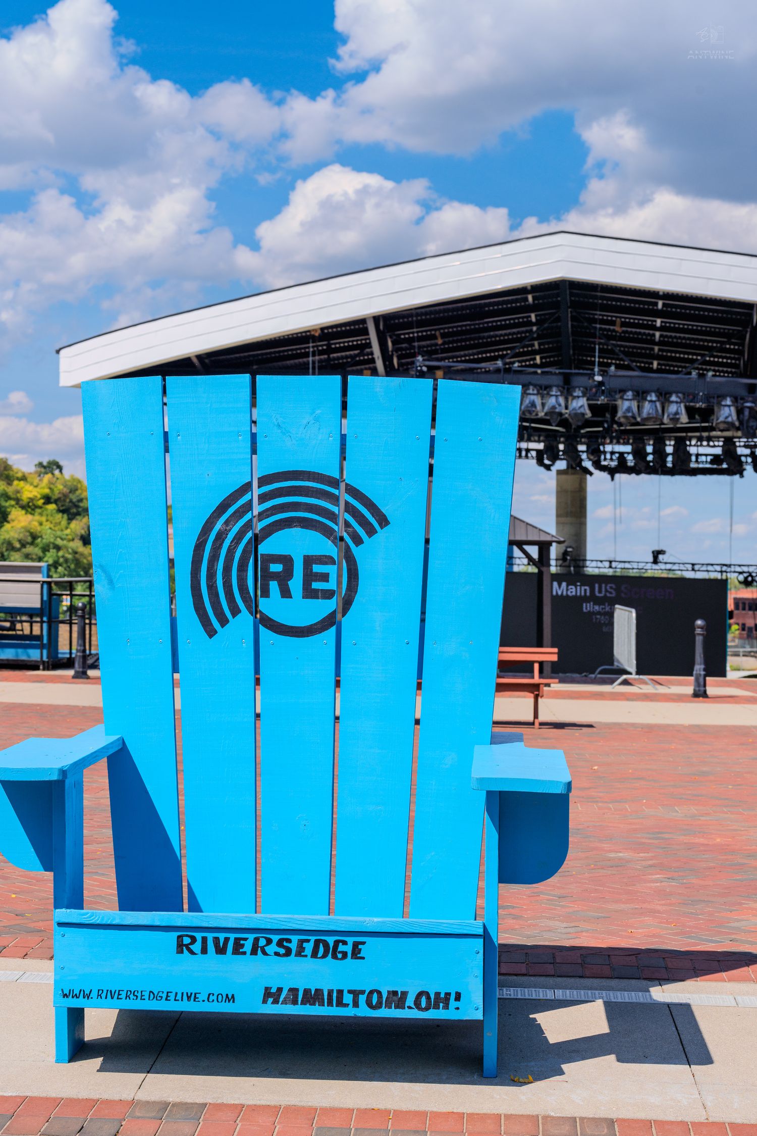 Giant blue Adirondack chair with RiversEdge logo at Hamilton, Ohio outdoor concert venue under blue sky.