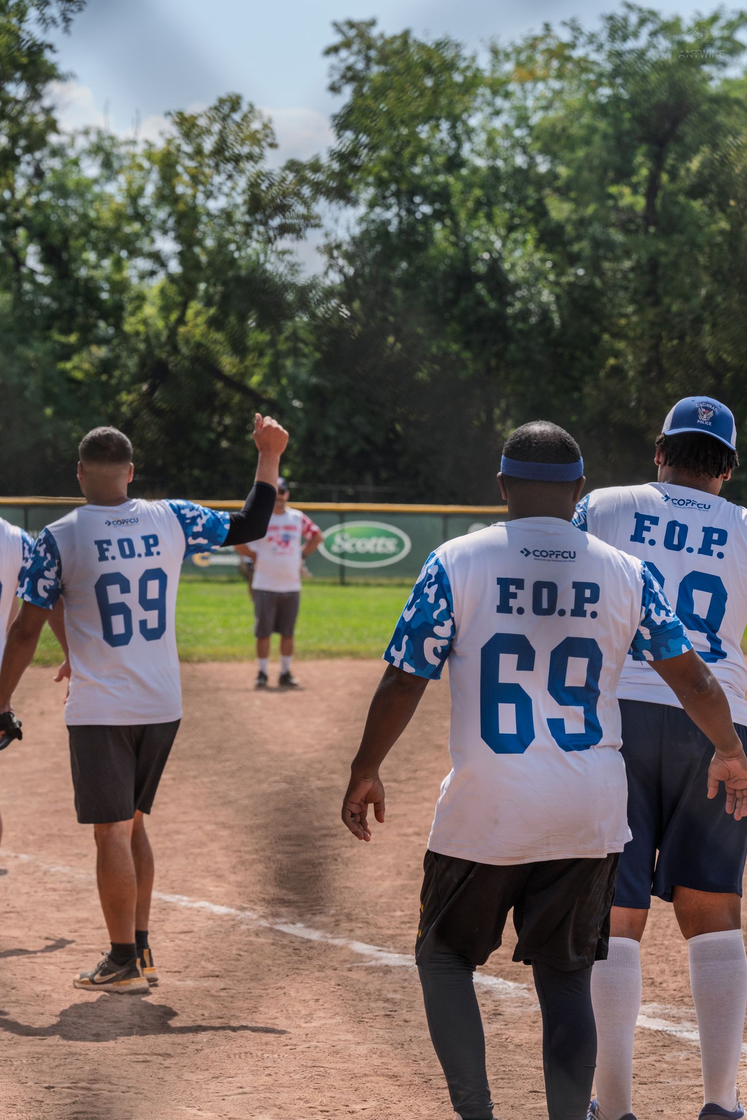 Baseball players in F.O.P. number 69 jerseys walk on a dirt field toward green trees on a sunny day.