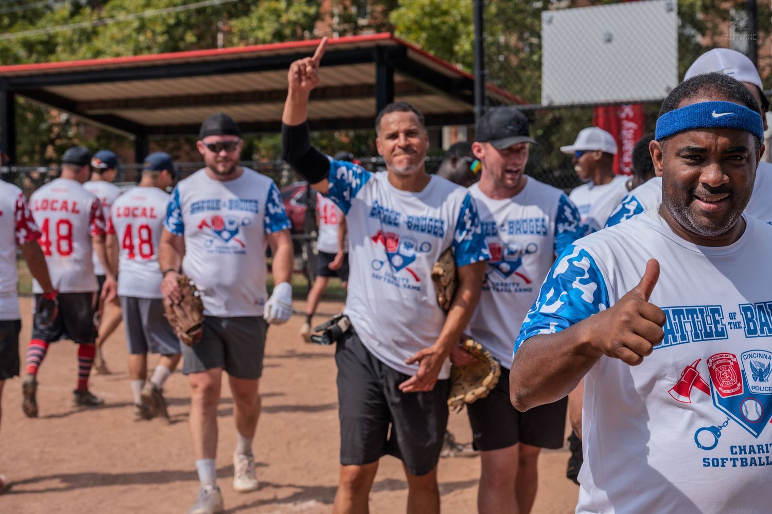 Team members in Battle of the Badges charity softball game jerseys cheer and give thumbs up on the field.