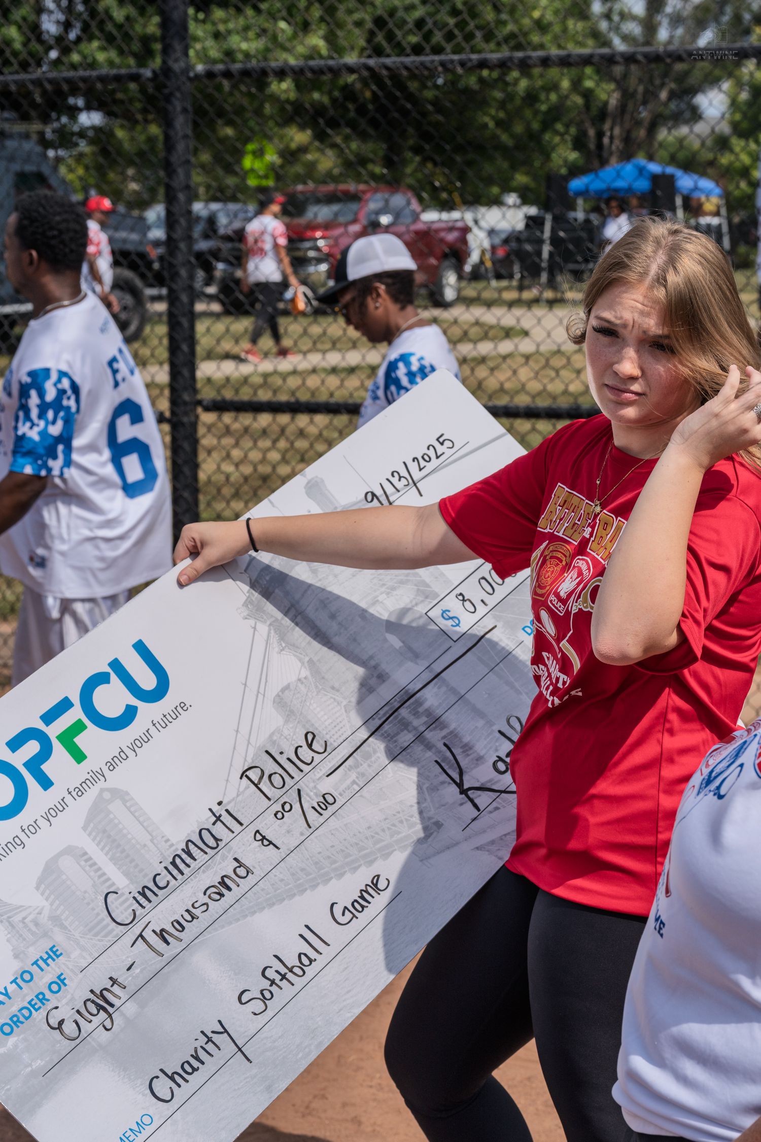 Young woman holds large ceremonial check for Cincinnati Police charity softball game sponsored by PFCU outdoors.