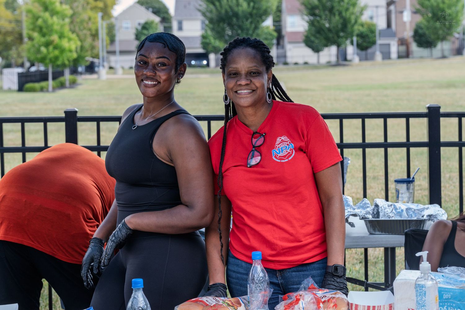Two women smile and pose together behind a food table at an outdoor charity softball game event.