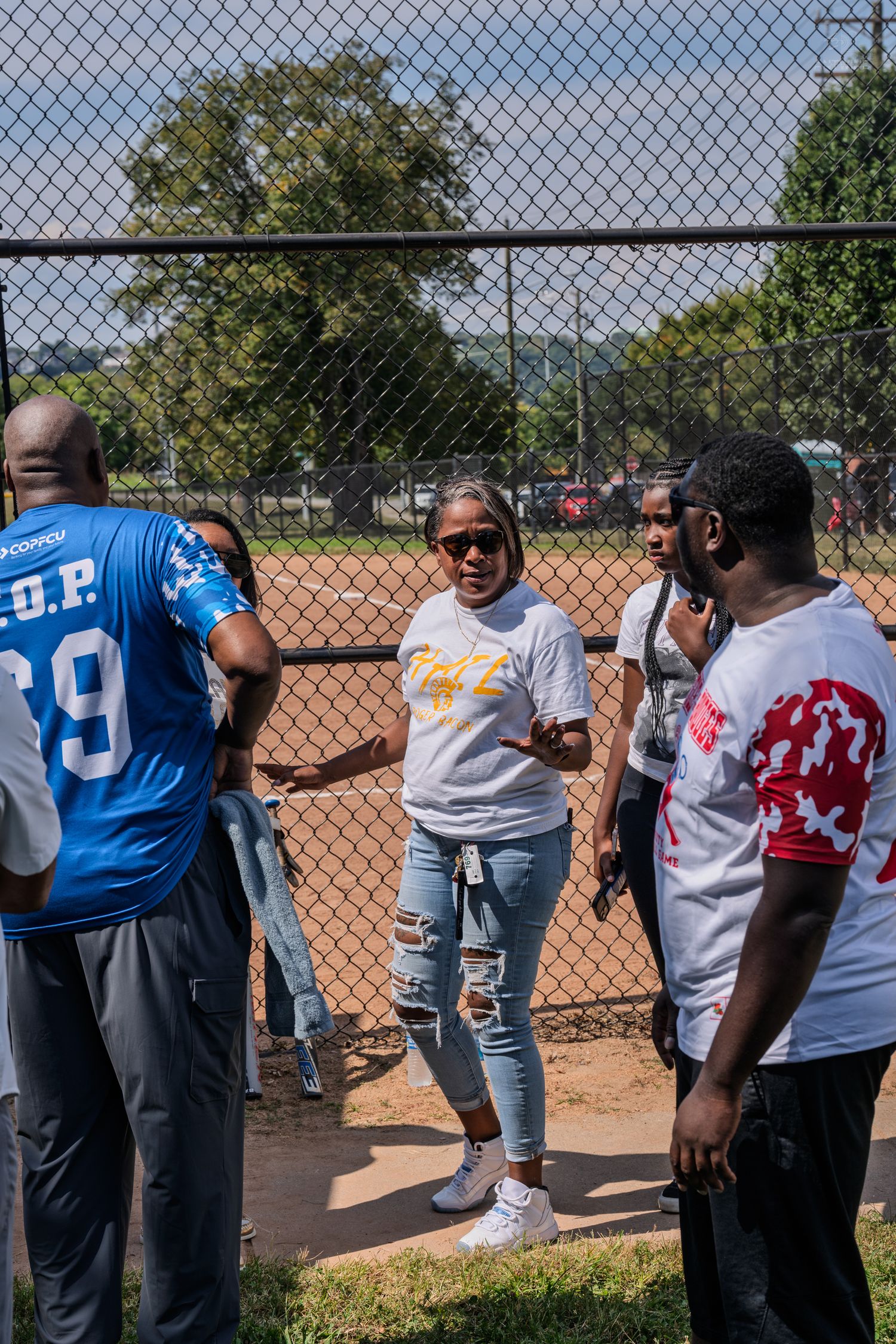Group of people in sports jerseys talking near a chain-link fence at an outdoor softball field on a sunny day.