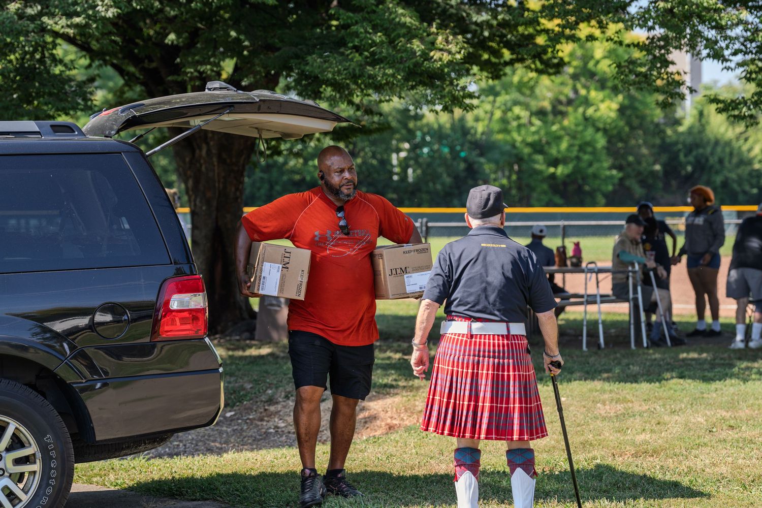Two men unloading boxes from an SUV outdoors, one wearing a red shirt and another in a traditional Scottish kilt.