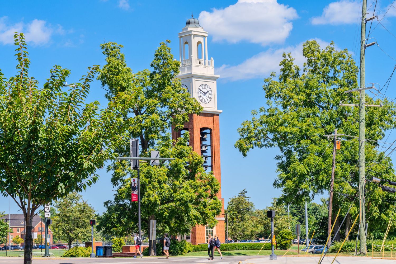 A red brick clock tower rises above lush green trees on a sunny university campus with a bright blue sky.