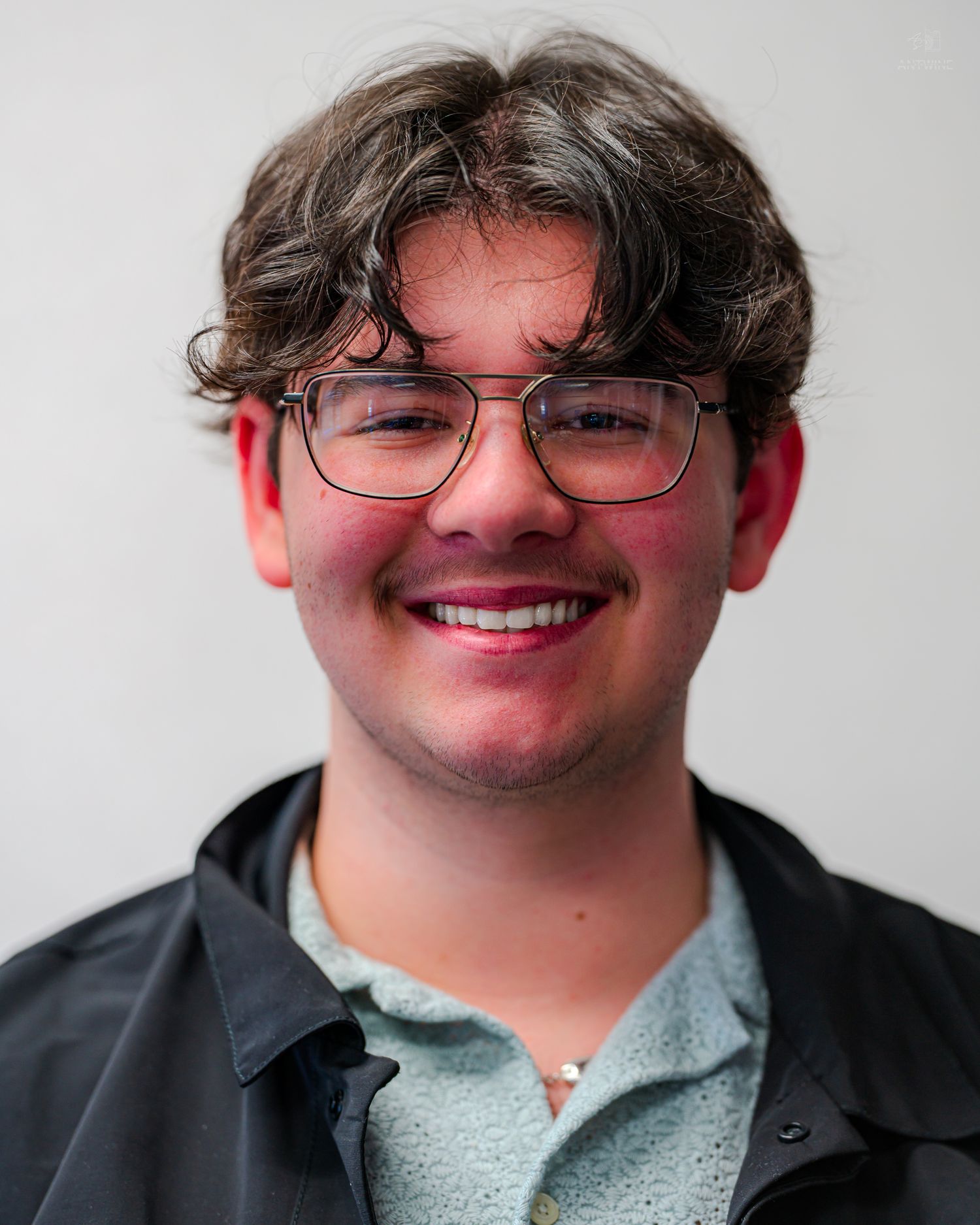Young man with curly hair and glasses smiling confidently in a light blue polo shirt and dark jacket.