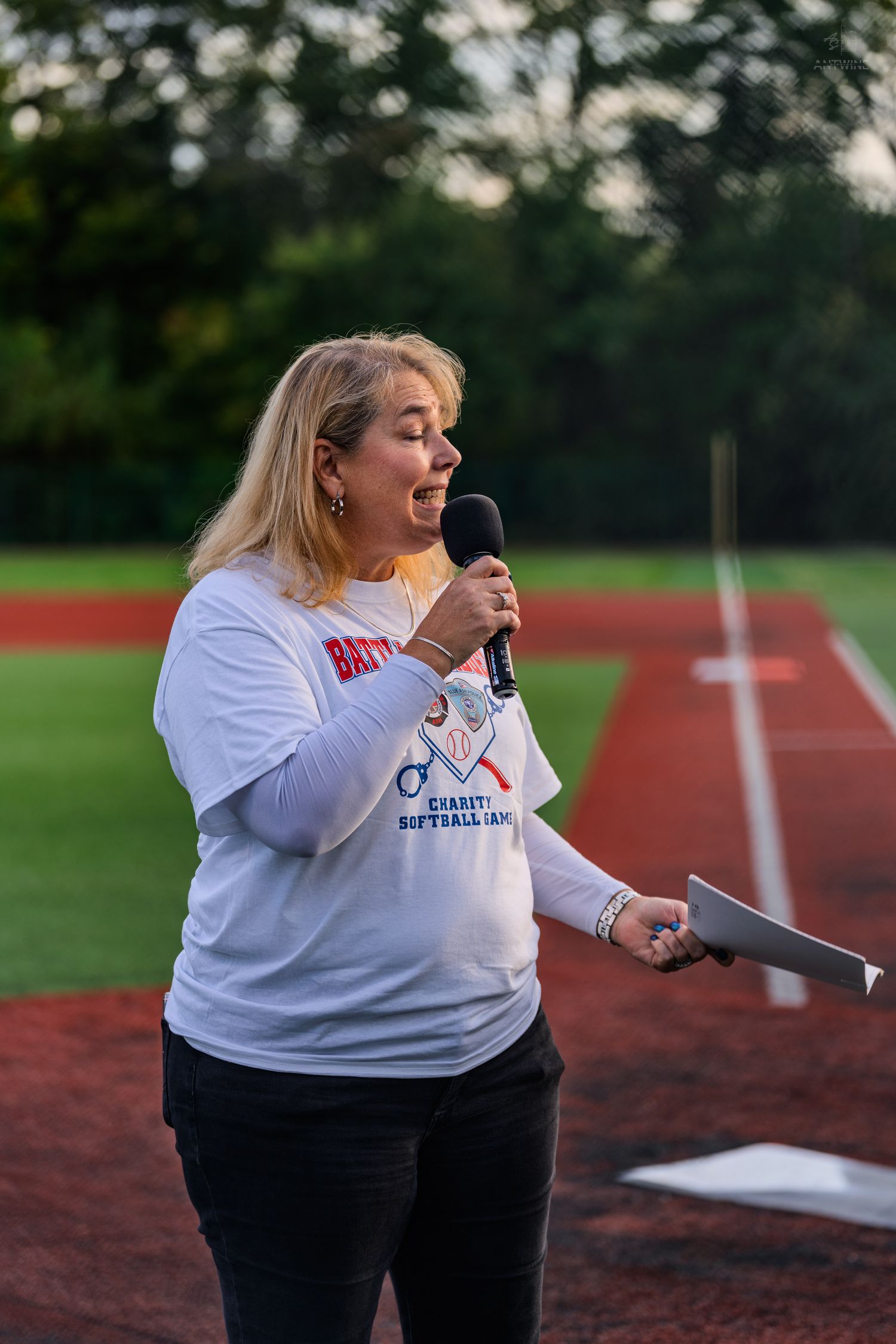 Woman speaking into microphone at charity softball game on baseball field at dusk.
