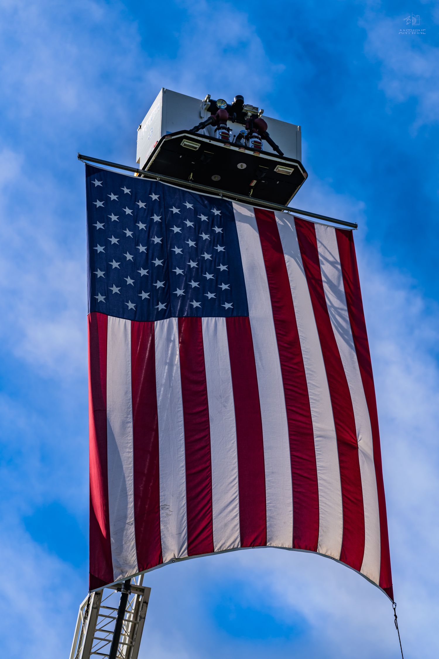 Large American flag hanging from a fire truck ladder against a blue sky, displaying stars and stripes prominently.