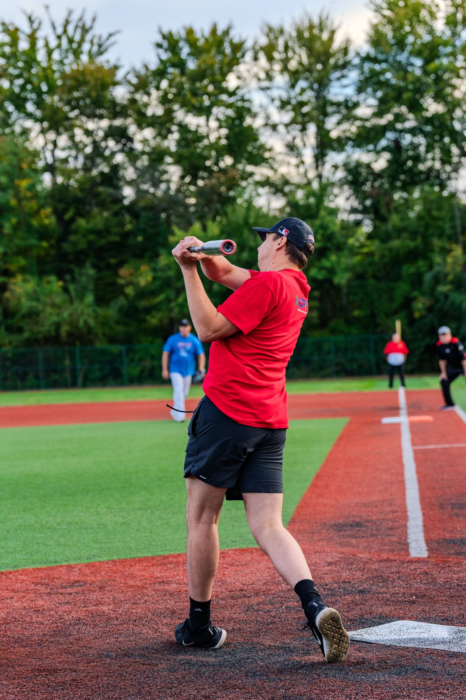A player in a red shirt swings a baseball bat at home plate during an outdoor softball game on a turf field.