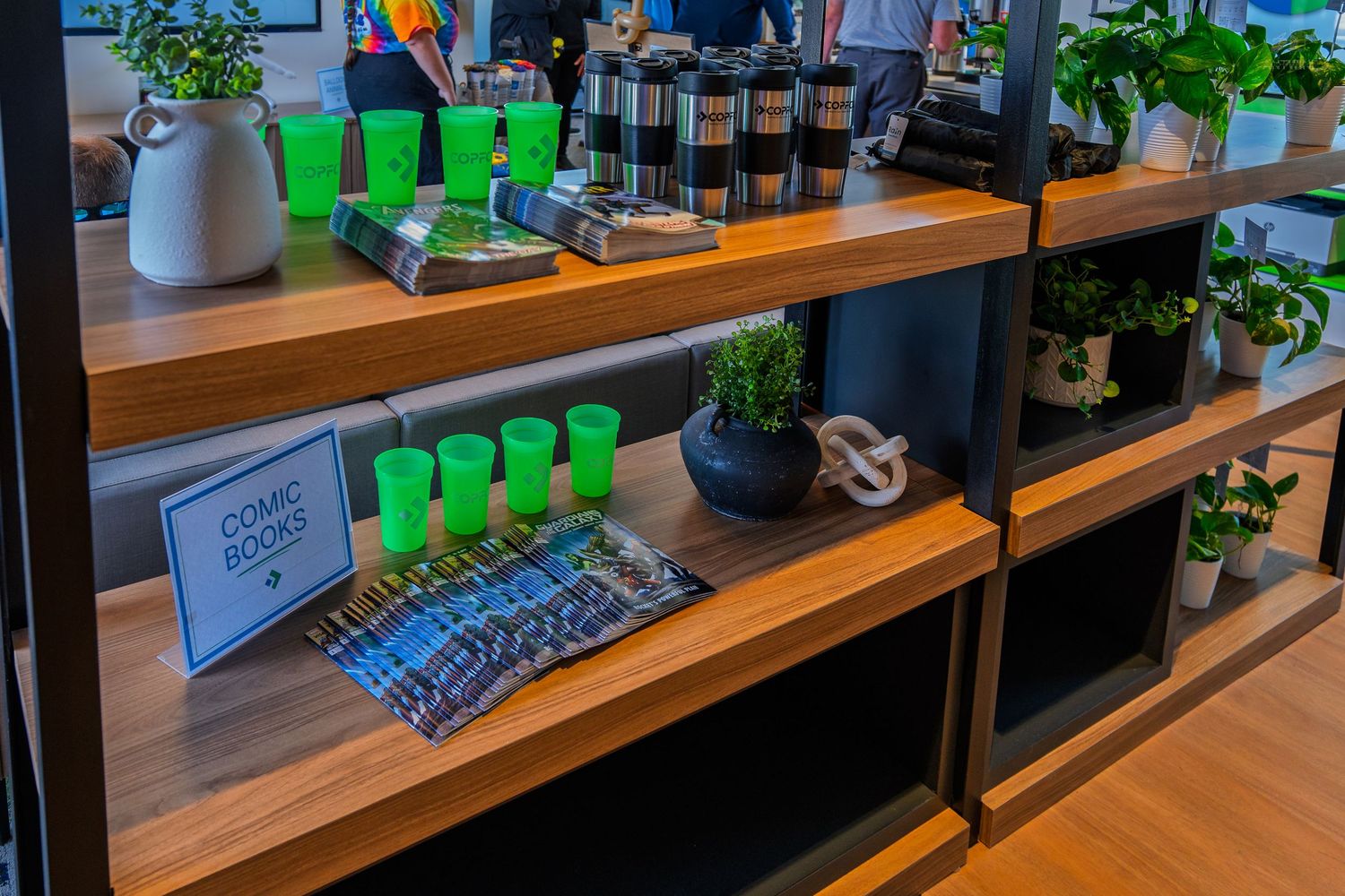 Wooden shelving unit displaying green cups, comic books, plants, and coffee accessories in a modern space.