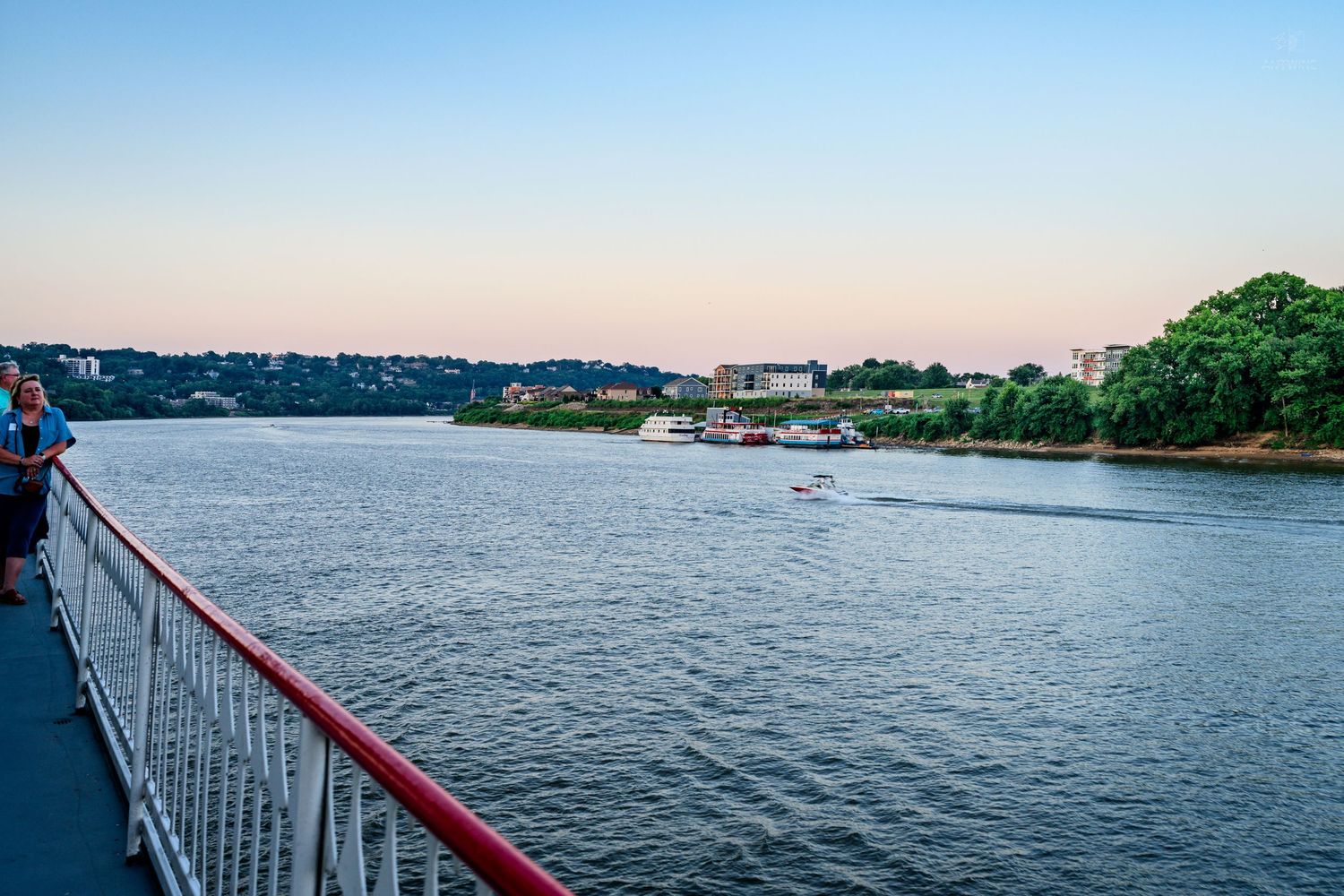 Scenic river view at dusk from a cruise ship deck with a red railing and distant cityscape.