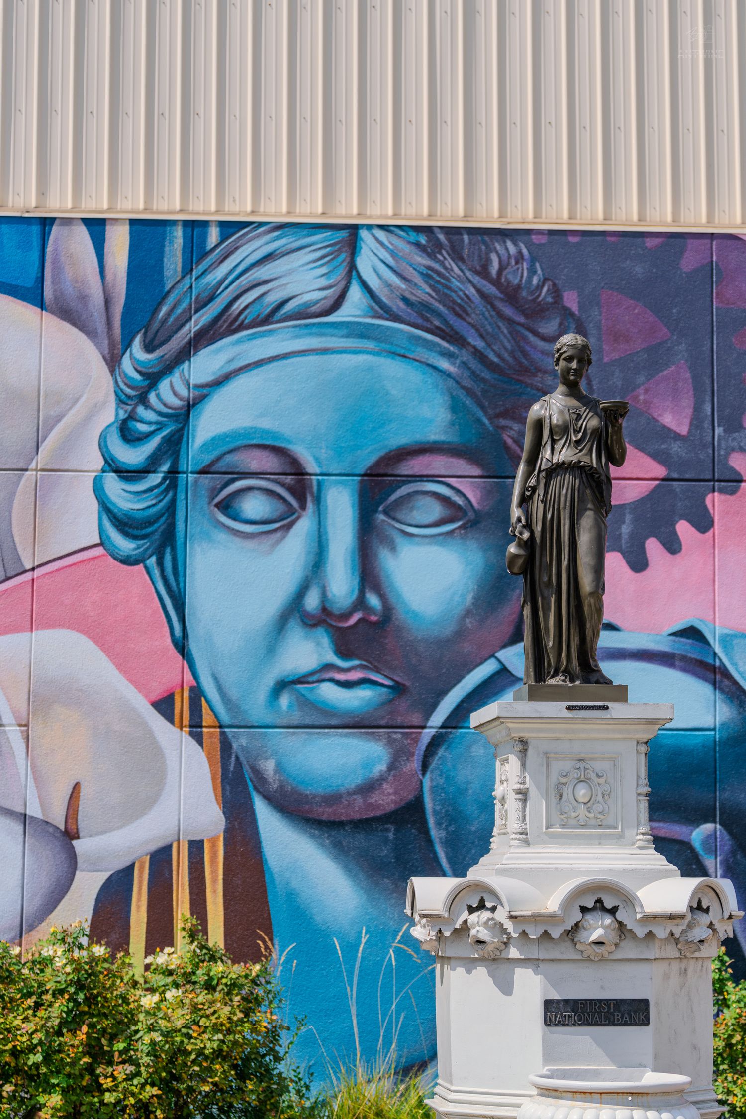 A bronze statue of a classical female figure stands before a vibrant blue mural of a woman's face on an urban wall.