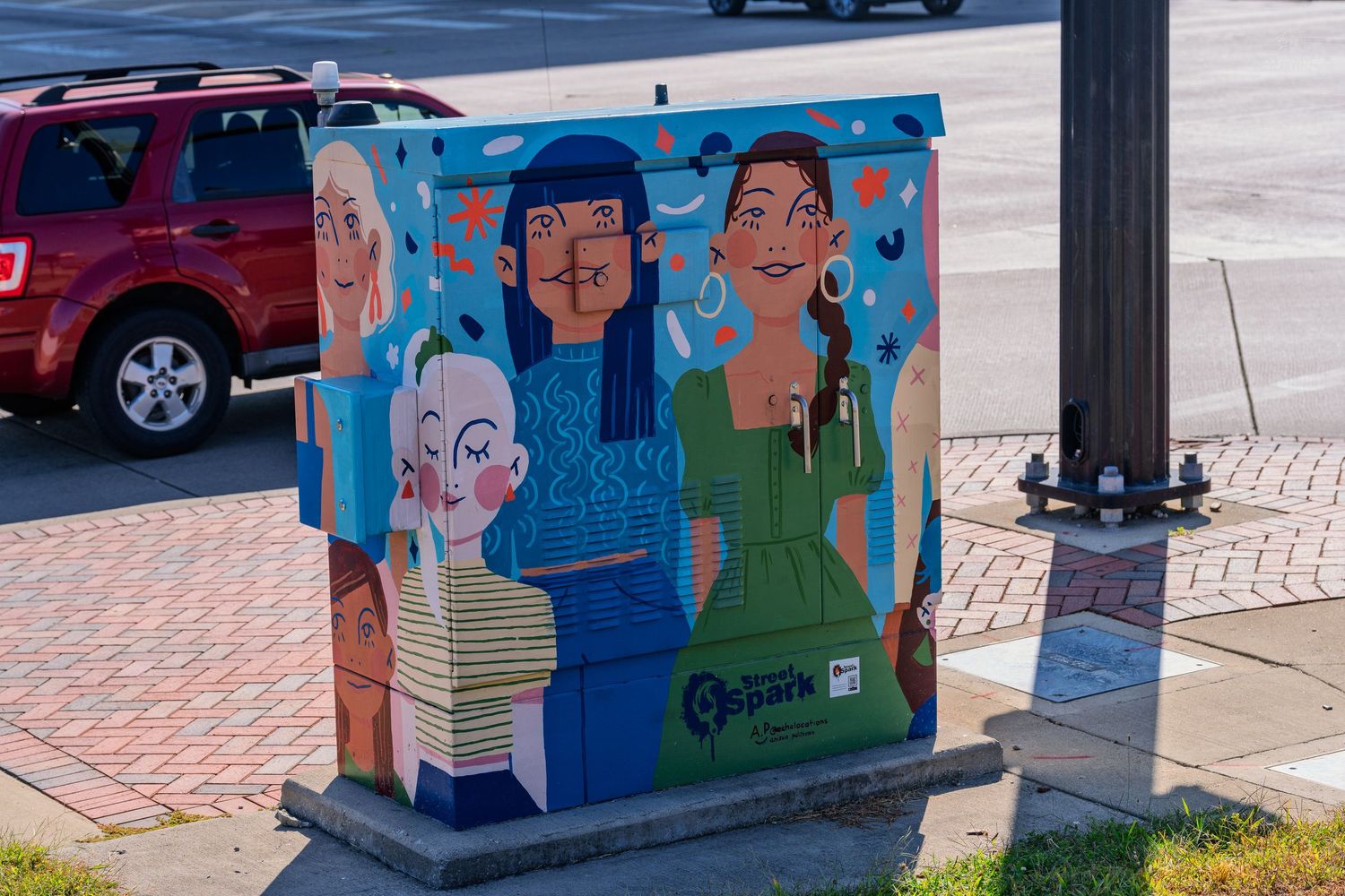 Colorful street art mural on utility box depicting four diverse women, located on a sunny urban sidewalk.